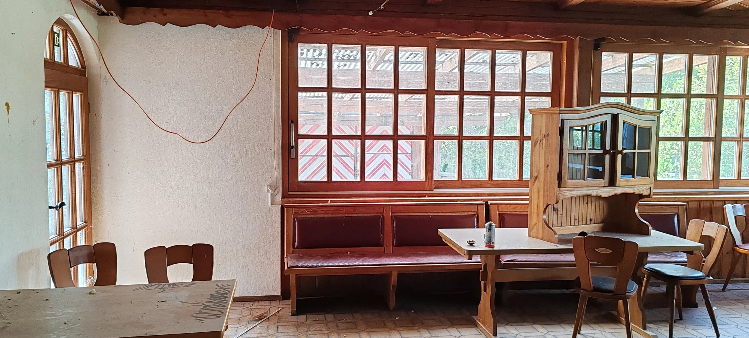 Innenraum eines Restaurants oder Cafés mit Holzmöbeln, großen Fenstern und sichtbarem Dach. Es gibt einen Tisch, Stühle, eine gepolsterte Sitzbank und eine Holzvitrine. Dekor ist einfach, mit einem roten und weißen Sonnenschirm draußen sichtbar.