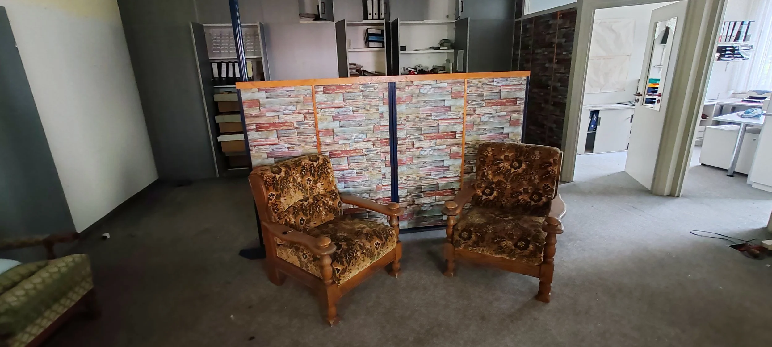 Zwei gemütliche Sessel mit Blümchenmuster in einem Raum, im Hintergrund eine Raumaufteilung mit Steinoptikwänden, ein Wasserautomat, und ein Bereich mit Aktenregalen und einem Büro.