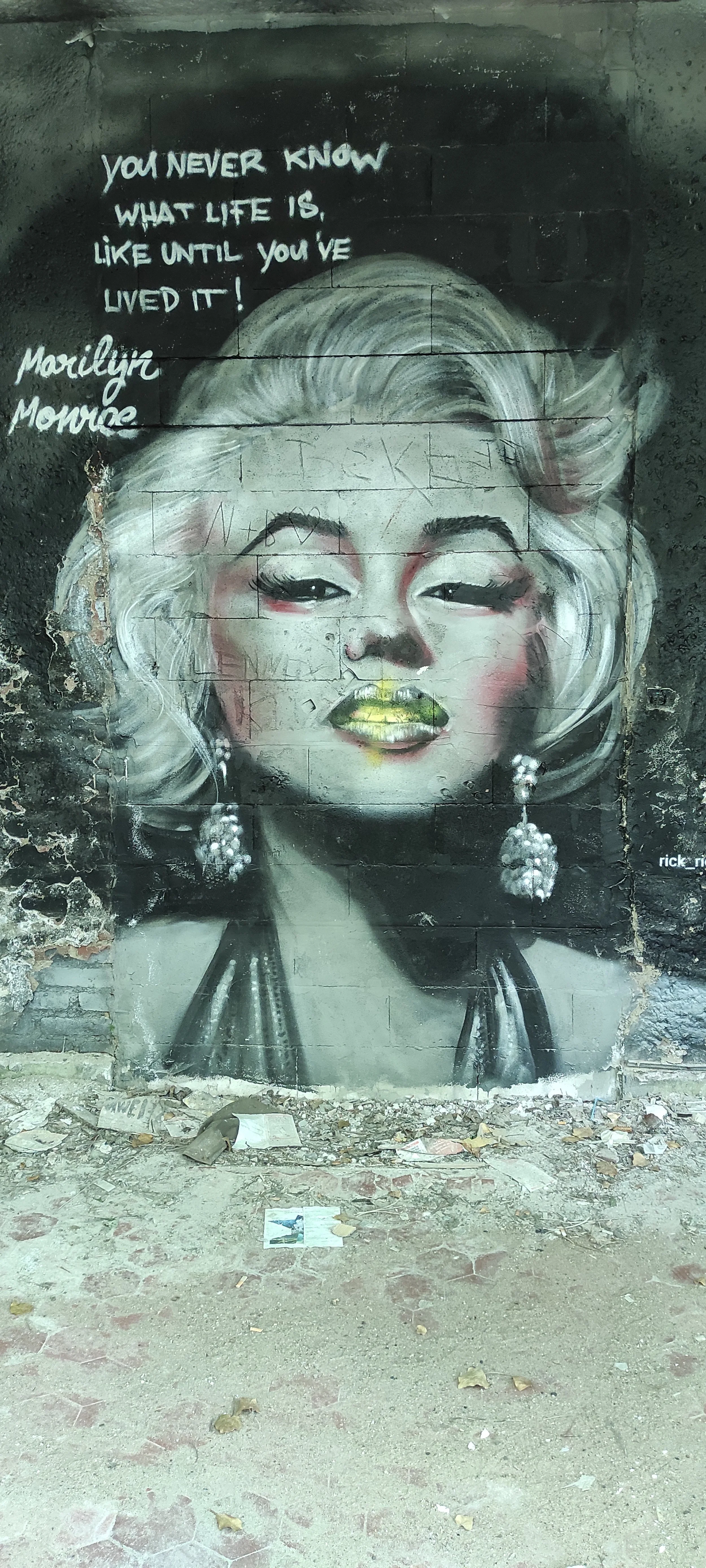 Wandgemälde einer Frau mit blondem Haar und großen Ohrringen, mit einem Zitat an der Wand: "You never know what life is, like until you've lived it!"