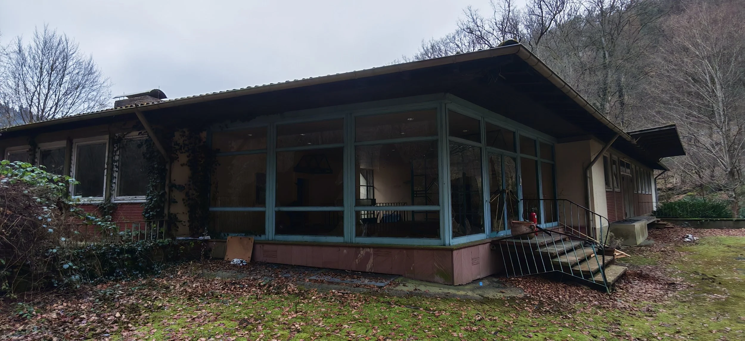 Ein verlassenes Haus mit großen Fenstern, umgeben von Bäumen und Laub im Herbst.