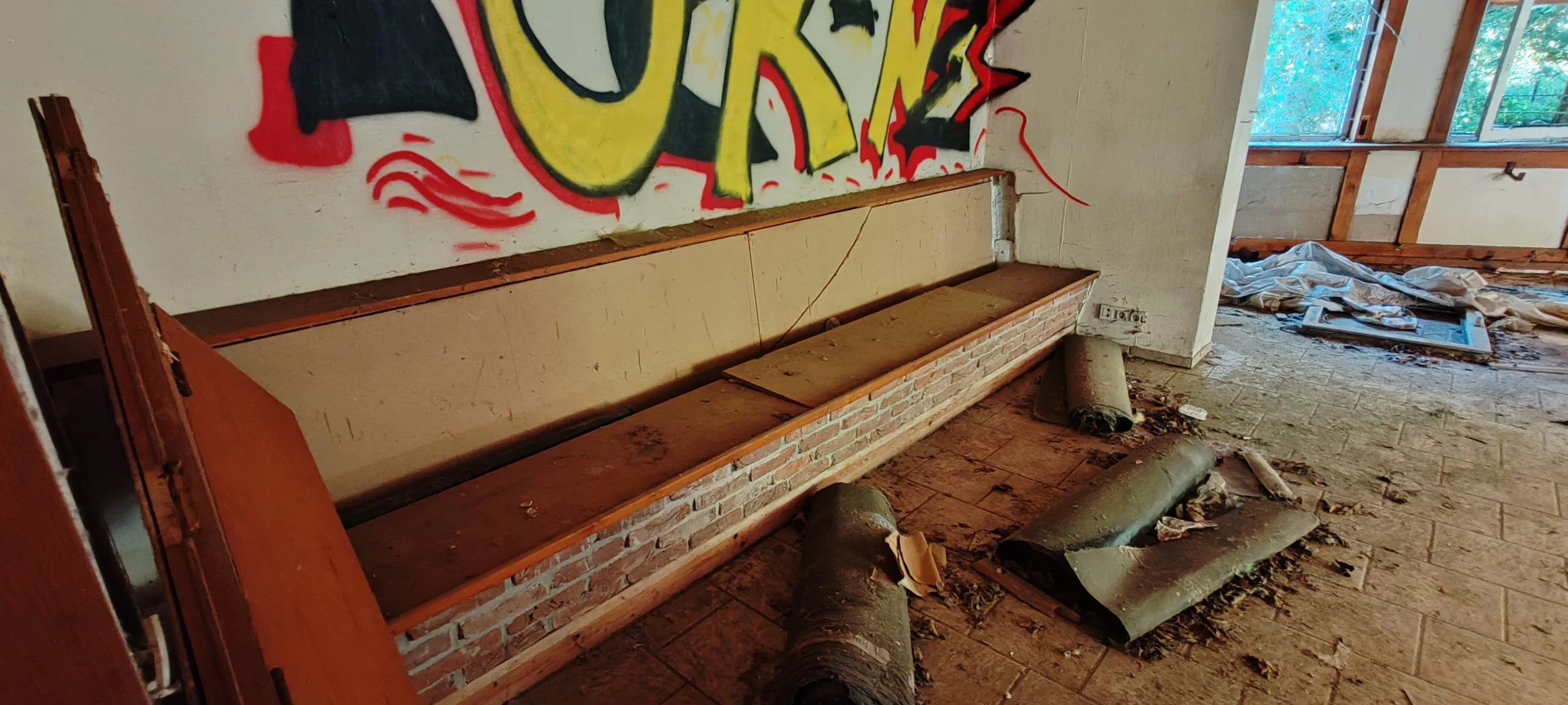 Innenraum eines verlassenen oder renovierungsbedürftigen Gebäudes mit einer alten Holzbank auf einer Ziegelmauer, zersplitterten Dachpfannen und herumliegendem Müll, darunter Holzteile und ein Rohr, sowie bunte Graffiti an der Wand, im Hintergrund Fe