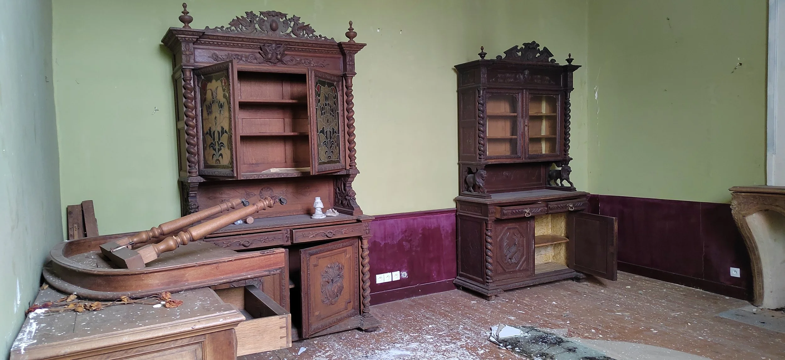 Ein Zimmer mit alten, kunstvoll geschnitzten Holzregalen und einem zerbrochenen Tisch, auf dem zwei heruntergefallene Gardinenstangen liegen. Der Boden ist staubig und mit Trümmern bedeckt, die Wände sind zwei-tone, grün und dunkelrot gestrichen.