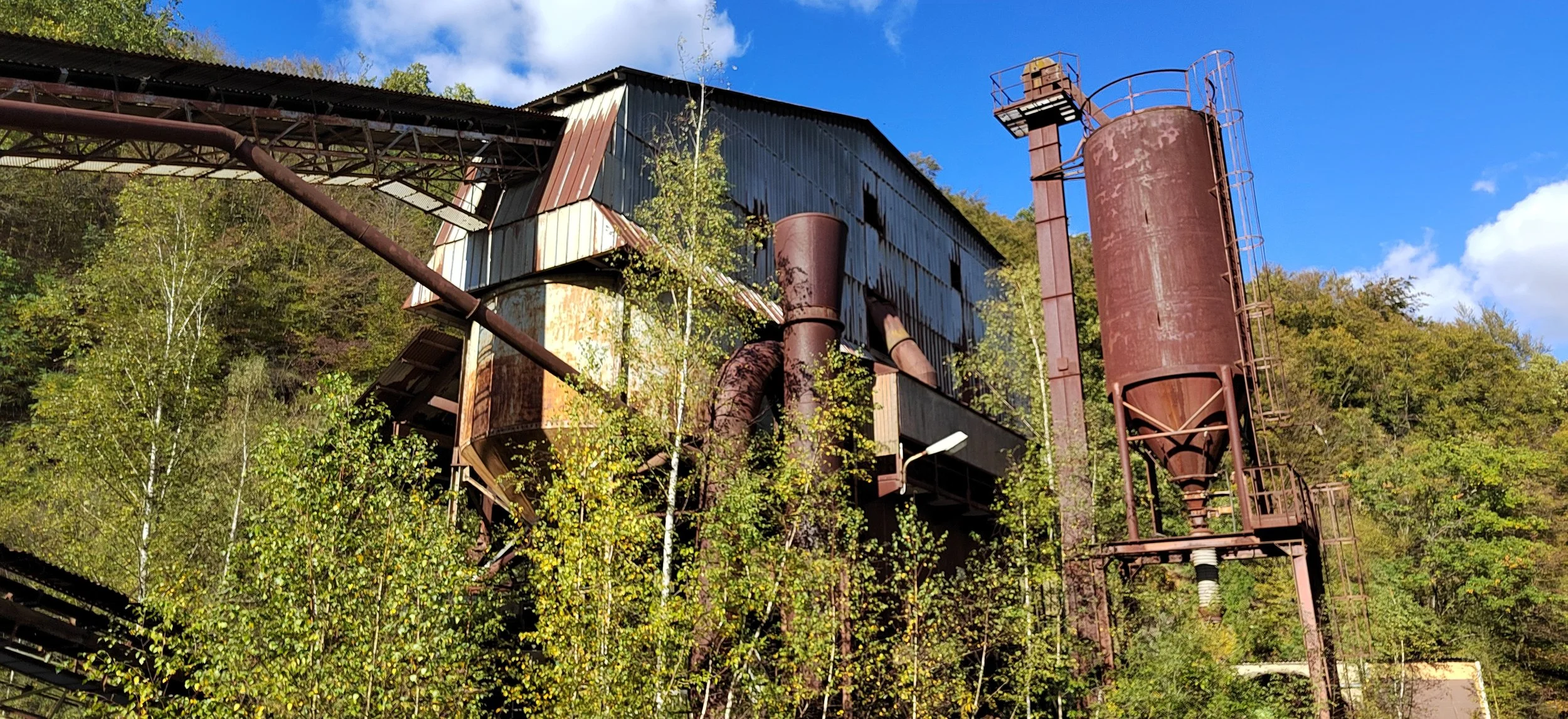Alte, verrostete Industrieanlage mit Silos und Rohren in einer bewaldeten Gegend unter einem blauen Himmel.
