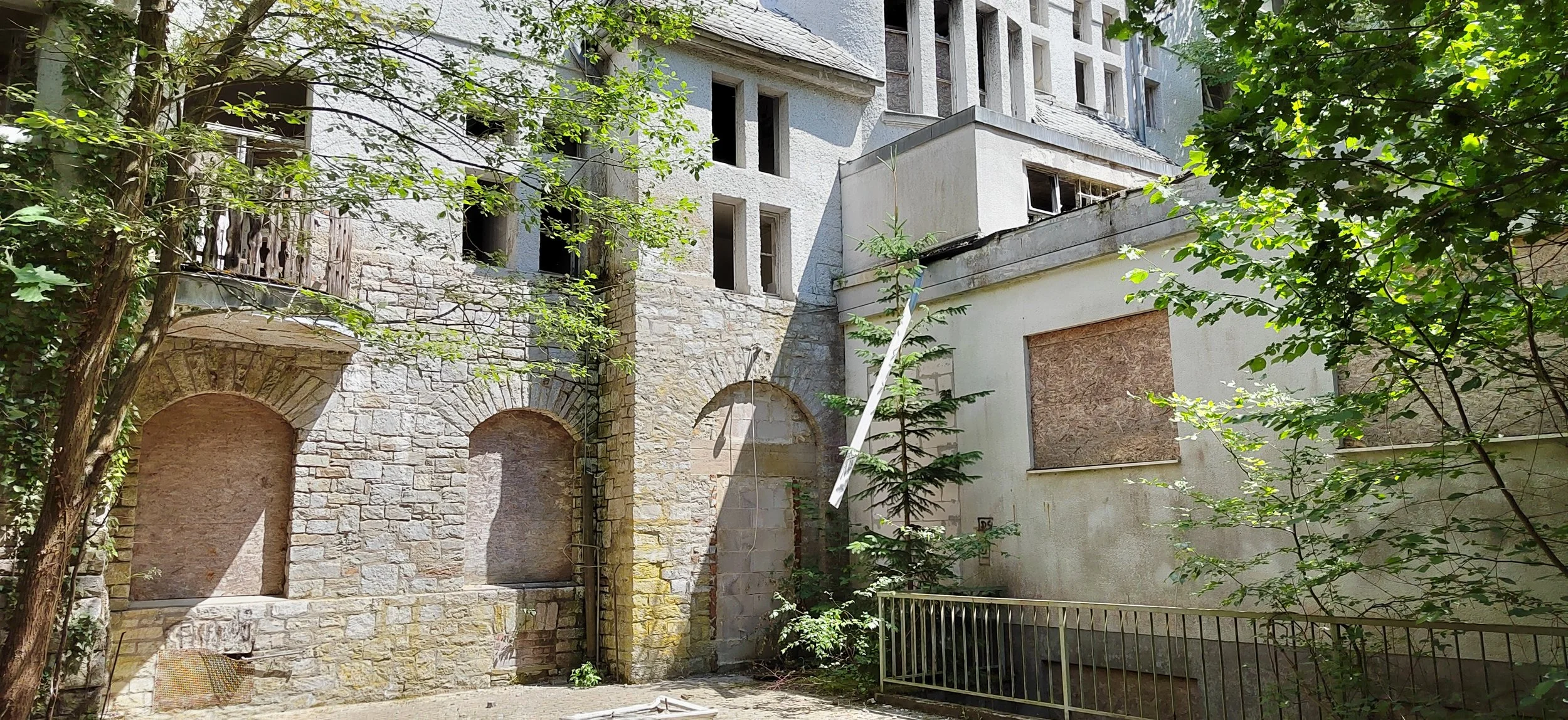 Ein altes, verfallenes Gebäude mit bröckelnder Steinmauer und leer stehenden Fenstern, umgeben von grünen Bäumen und Vegetation, in einem Innenhof.