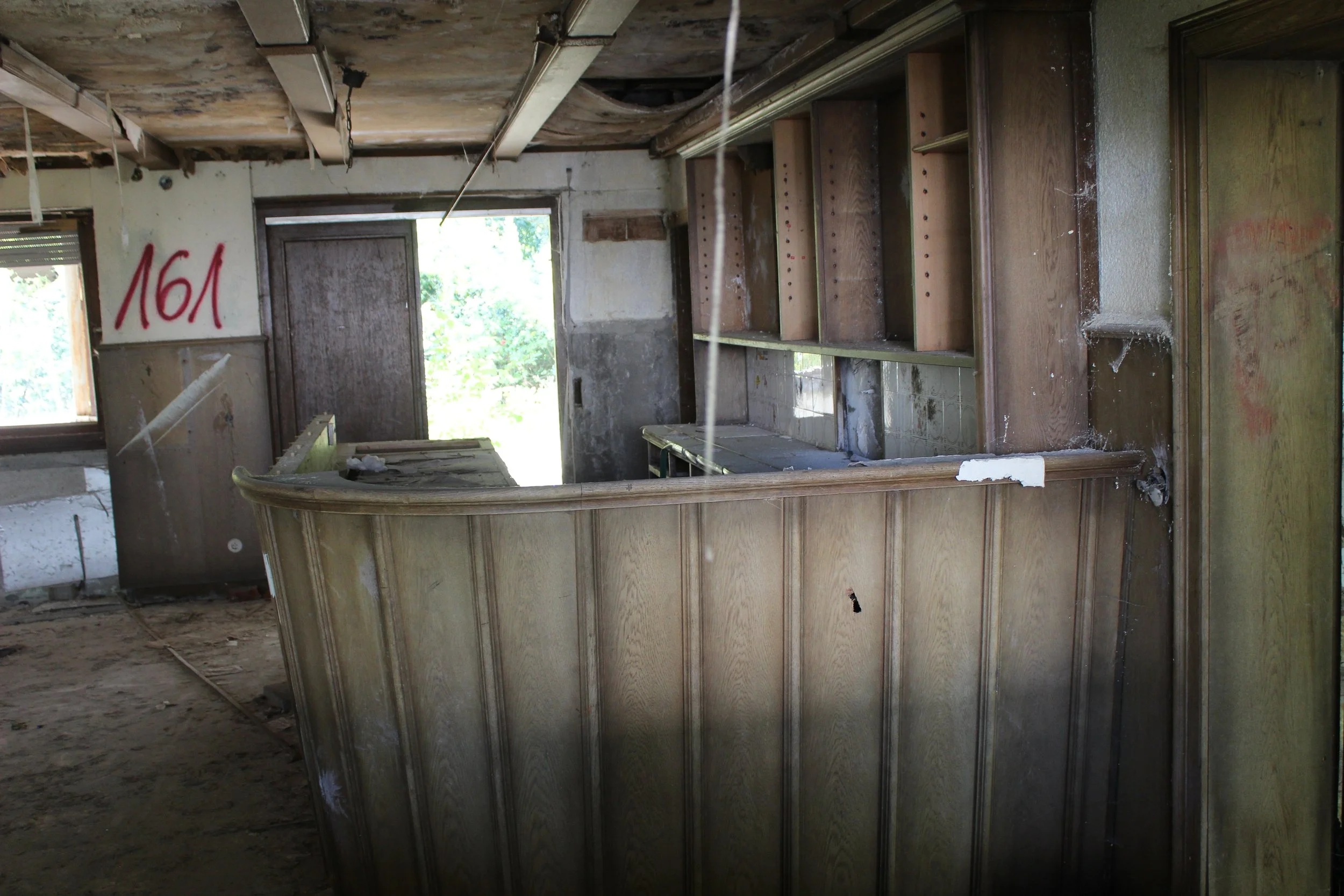 Innenansicht eines verfallenen, leeren Raumes mit Holztheke, offenen Regalen und kaputtem Boden. Fenster lässt Tageslicht herein, auf einer Wand befindet sich rote Graffiti '161'.