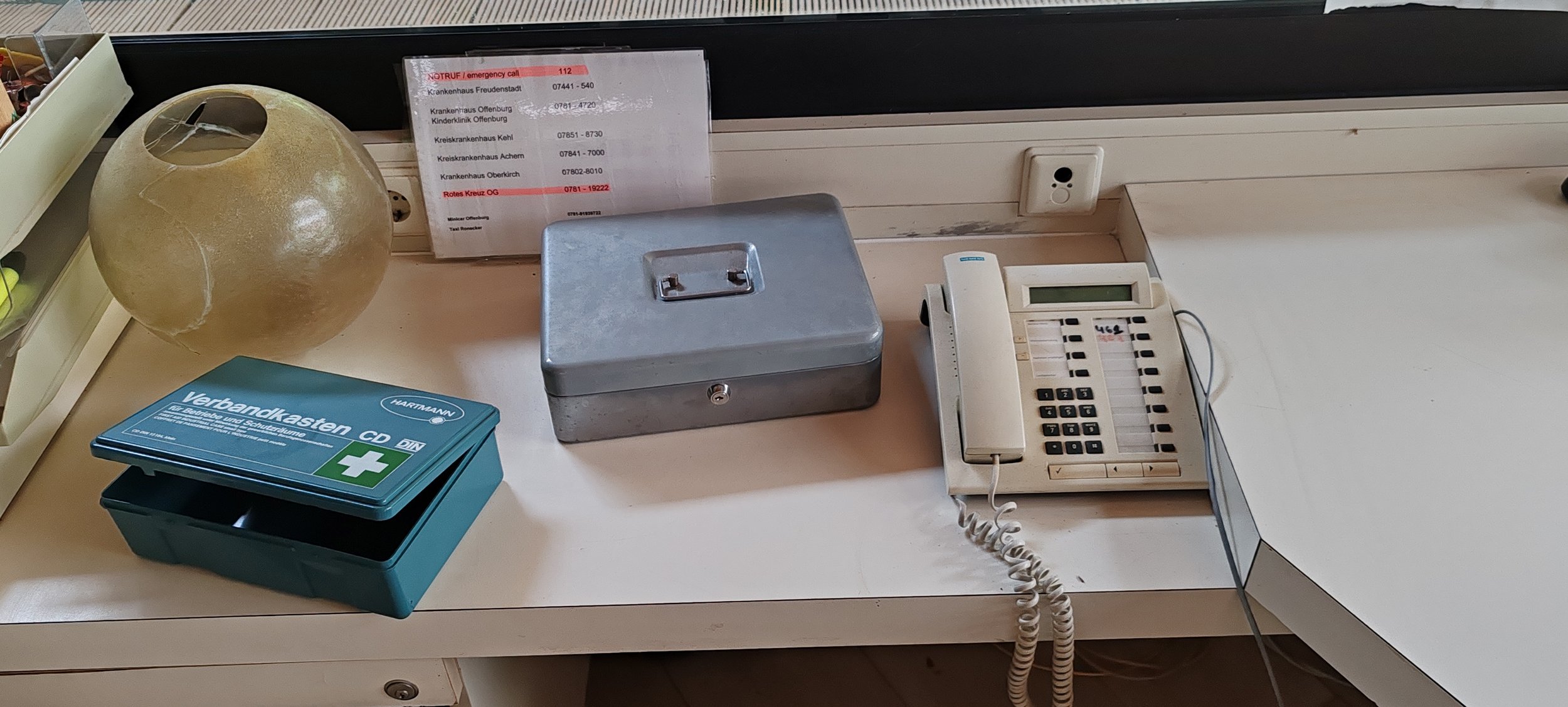 Büro- oder Empfangstresen mit einem Telefon, einem Metal-Aktenkoffer, einem Briefkasten für Verbandskarten und einer roten Telefonliste an der Wand.