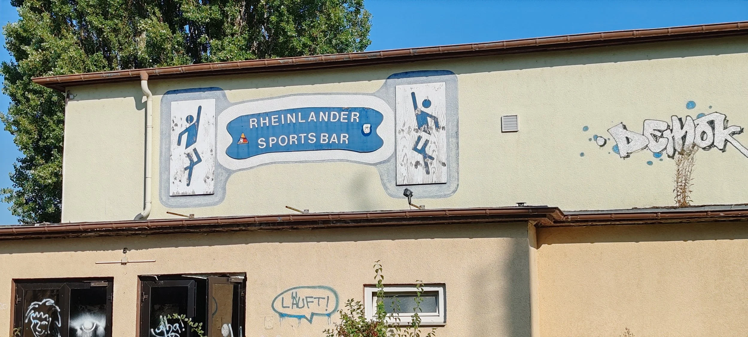 Grafitti und ein Schild an einer Hauswand, das den Rheinländer Sports Bar anzeigt, mit Sport- und Freizeitsymbolen sowie weiteren Graffiti-Elementen und einem Baum.