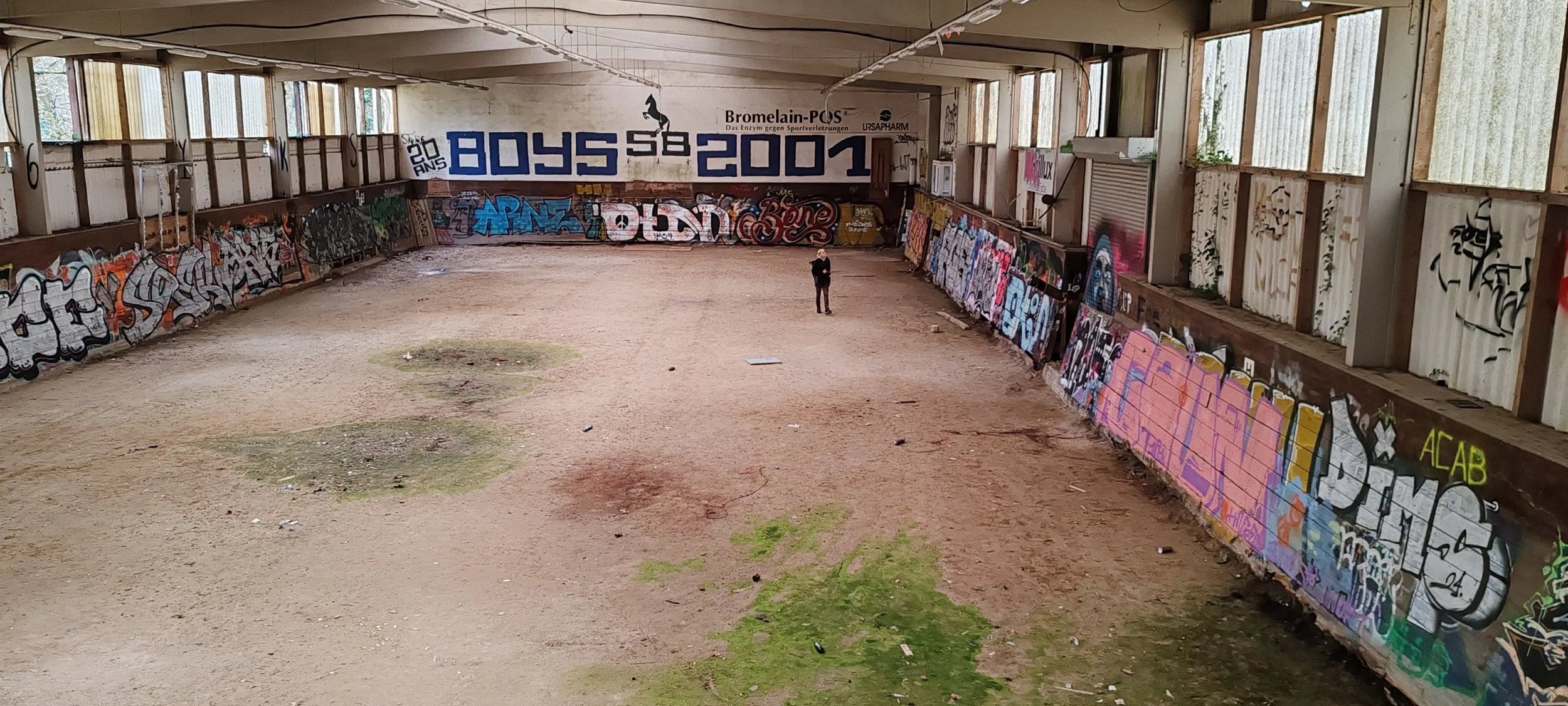 Ein verlassener Hallenboden mit Graffiti an den Wänden, eine einzelne Person steht in der Mitte des Raumes, der Raum ist mit Sonnenlicht durch Fenster an den oberen Wänden erhellt.