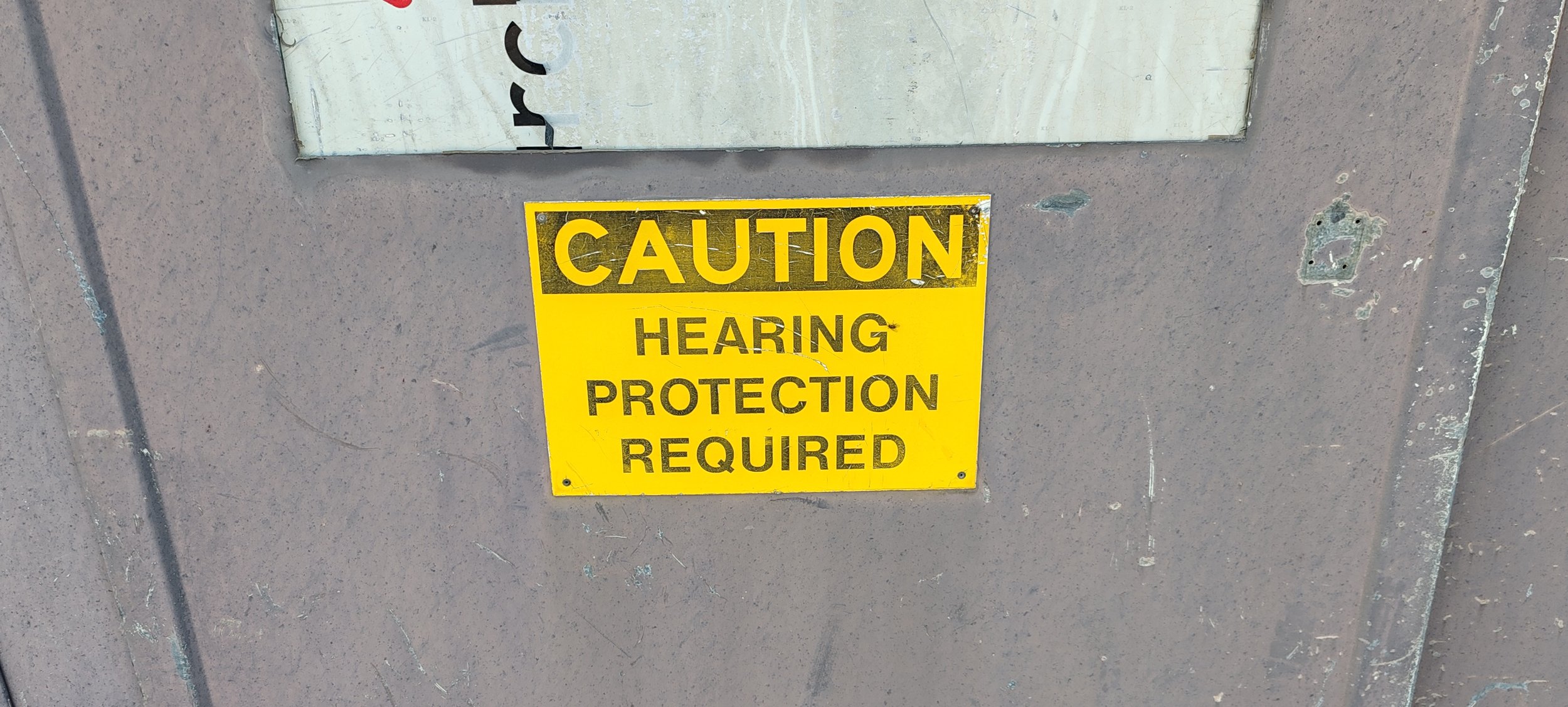Gelber Warnhinweis auf einer Wand, mit schwarzem Text: "Vorsicht, Gehörschutz erforderlich".