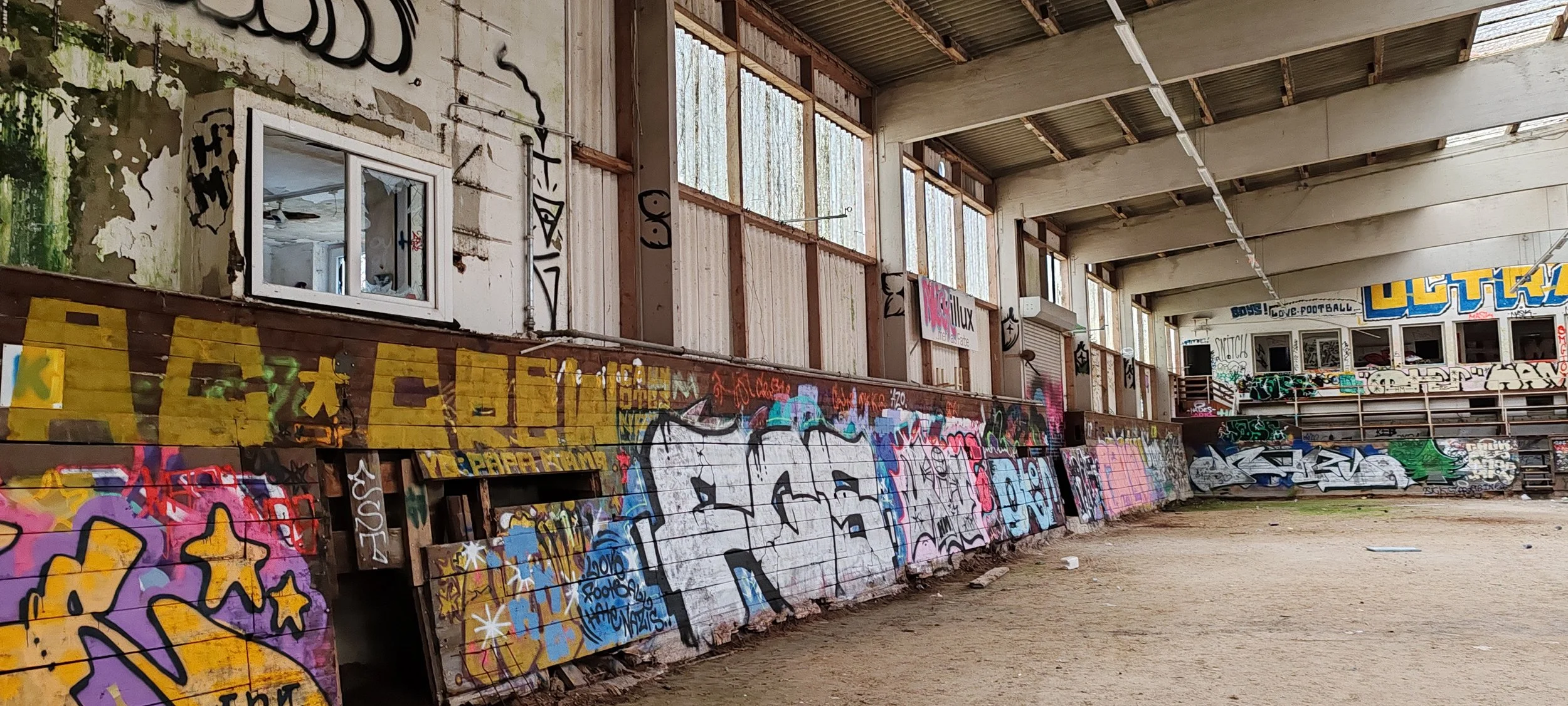 Innenansicht eines alten, verlassenen Sports Stadions mit grauer, verfallener Wand und mehreren bunten Graffiti. Es gibt eine kleine Fensteröffnung und eine Tribüne im Hintergrund.