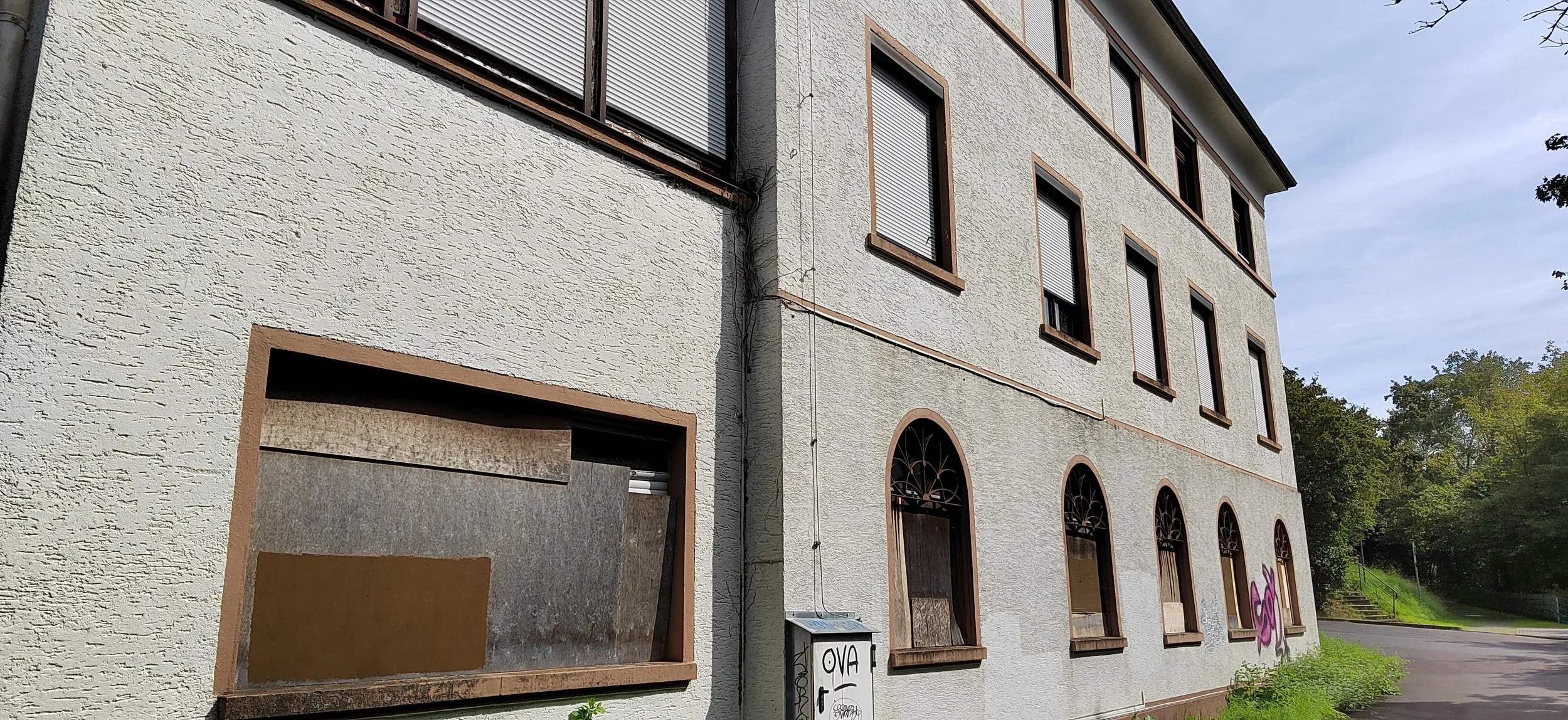 Ein altes mehrstöckiges Gebäude mit mehreren verschlossenen Fenstern, einige mit Rollläden, an einer Seite befindet sich Graffiti. Es gibt eine kleine Tür mit Glas an der unteren linken Seite und Grünflächen daneben.