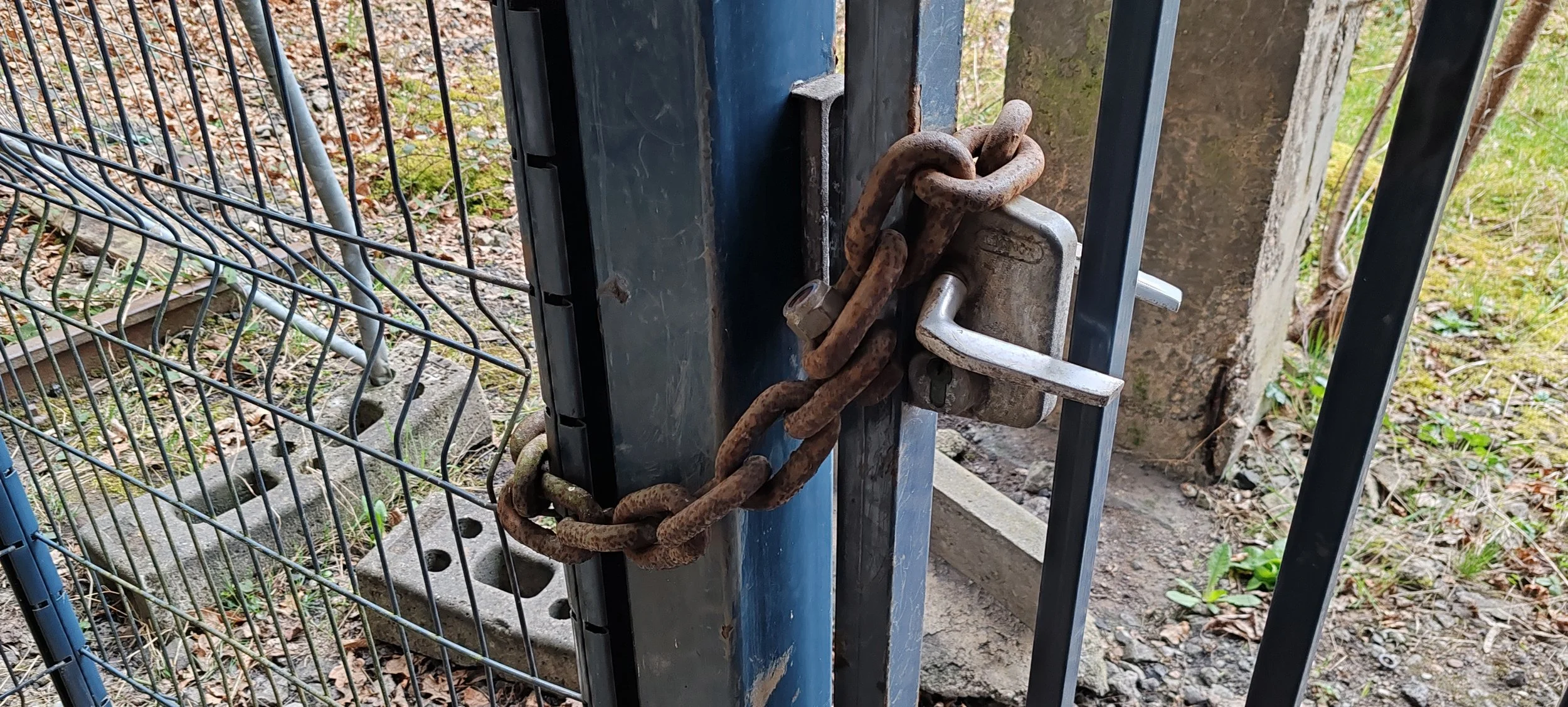 Verrostete Kette und Schloss sichern einen Torflügel vor unbefugtem Zutritt in einem Garten oder Außenbereich, mit Erde, Blättern und Pflanzen im Hintergrund.