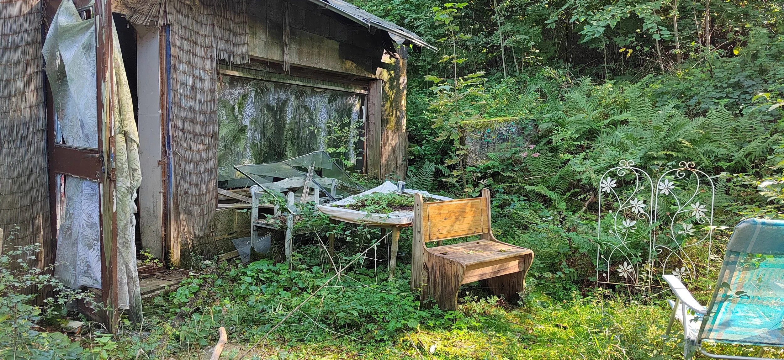 Ein verfallenes Gartenhaus umgeben von überwucherten Pflanzen, mit einem alten Stuhl und einer Tischplatte im Grünen, sowie Gartenmöbeln und Dekorationen im Außenbereich.