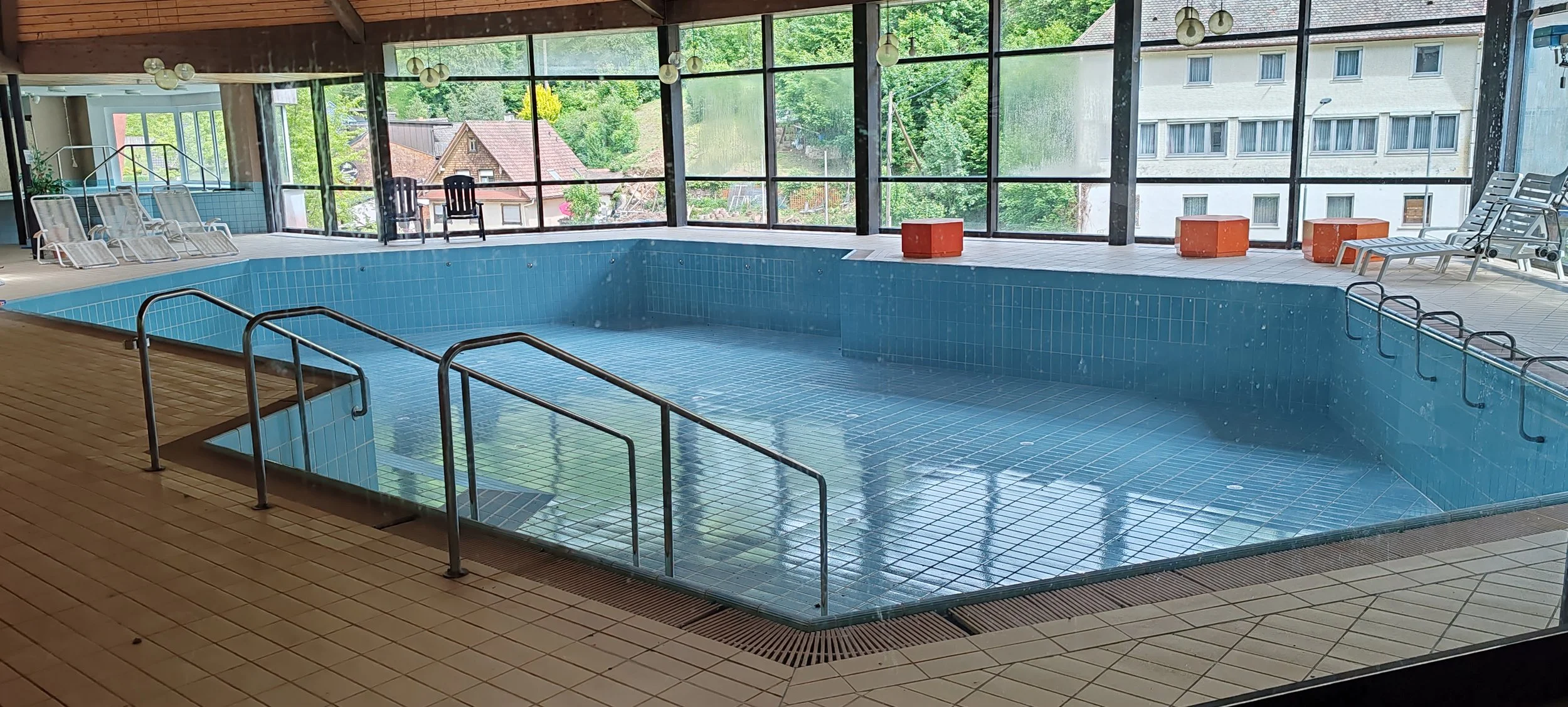 Innenansicht eines leeren Schwimmbads mit blauer Fliesen und Panorama Fenster, Stühle und Tische im Hintergrund, Blick auf grüne Bäume und Nachbarhäuser.