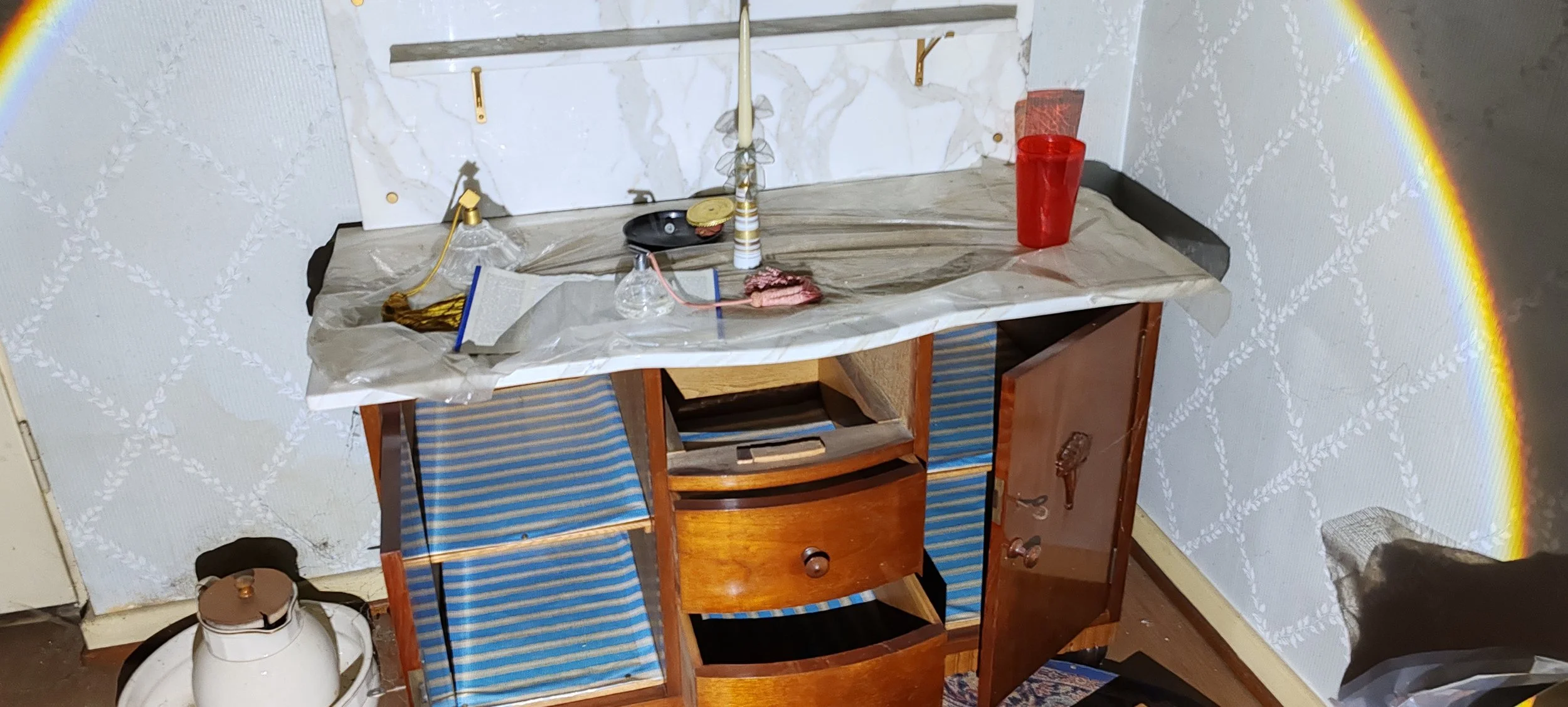 Ein alter, dreckiger Küchentisch mit einer Geschenkverpackung, Kerzen, einem roten Glas und anderen Gegenständen. Gegenüber ist ein schrank mit abstehenden Türen. Der Raum wirkt ungepflegt.