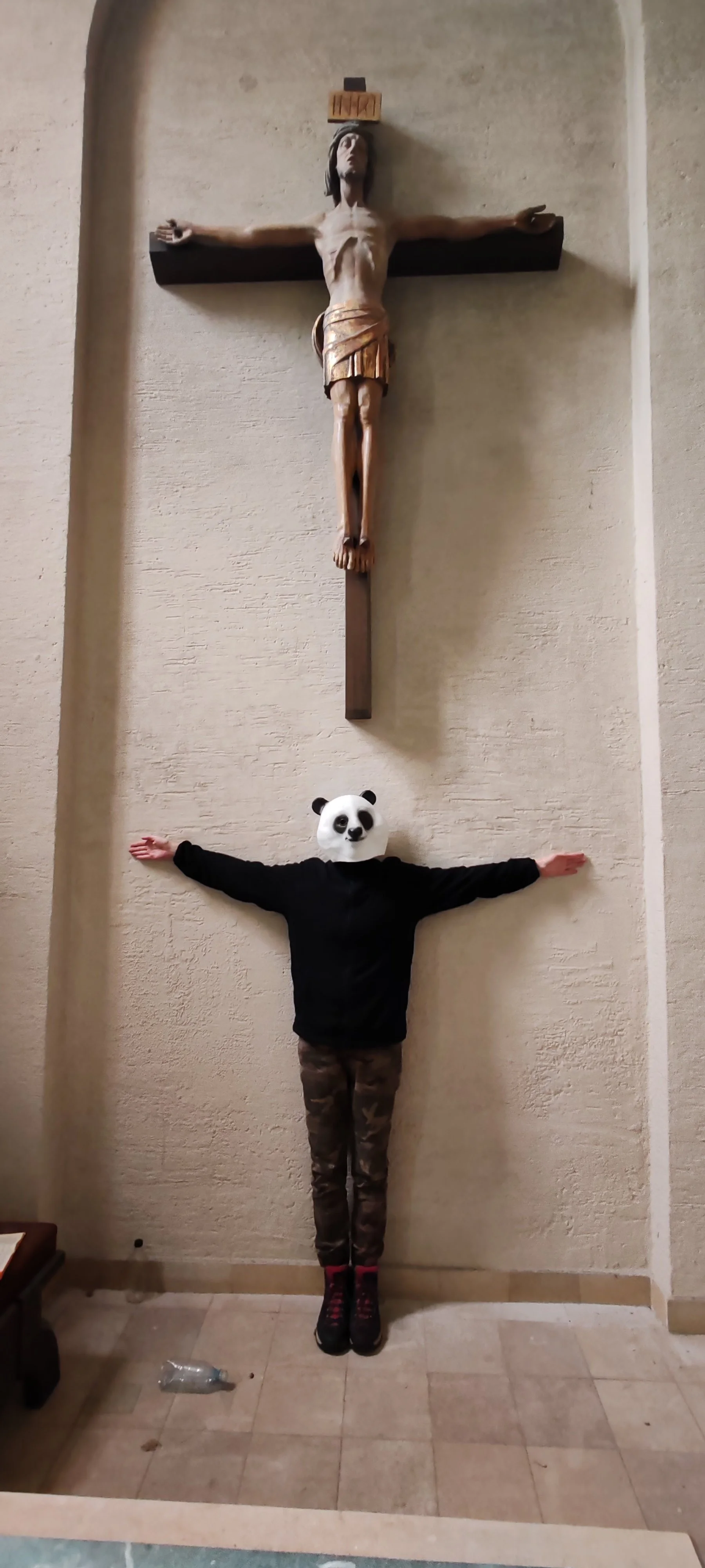 Person mit Pandakopf steht vor einer Wand, die zwei hängende Figuren zeigt: oben ein Christuskorpus und darunter eine Person mit Pandakopf, ausgestreckten Armen und camouflagemusternder Hose.