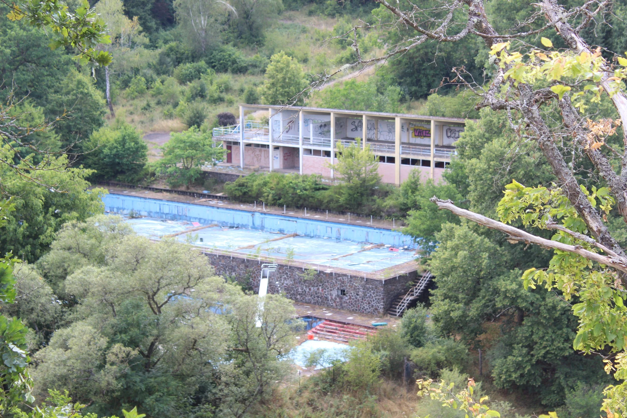 Verlassenes Schwimmbad im Grünen mit umgebender Vegetation und einem modernen Gebäude im Hintergrund.