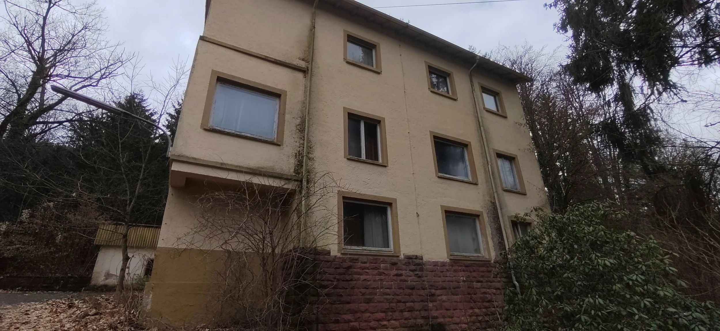 Mehrstöckiges altes Wohngebäude mit vier Fenstern pro Etage, umgeben von Bäumen und Sträuchern, bei bewölktem Himmel.