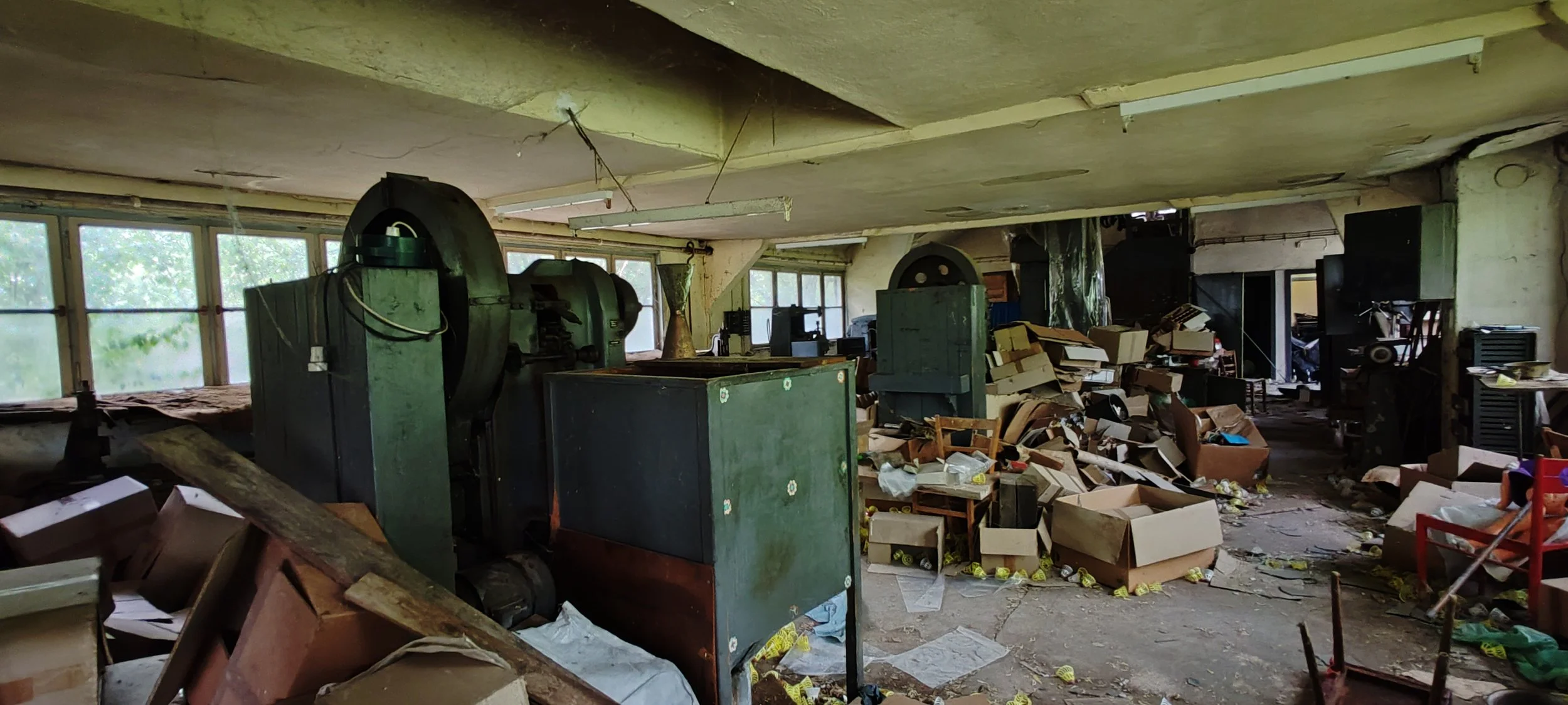 Ein verlassener Raum mit alten Druckmaschinen, verstreutem Verpackungsmaterial und unordentlichen Gegenständen auf dem Boden, in einem lichtdurchfluteten Gebäude.