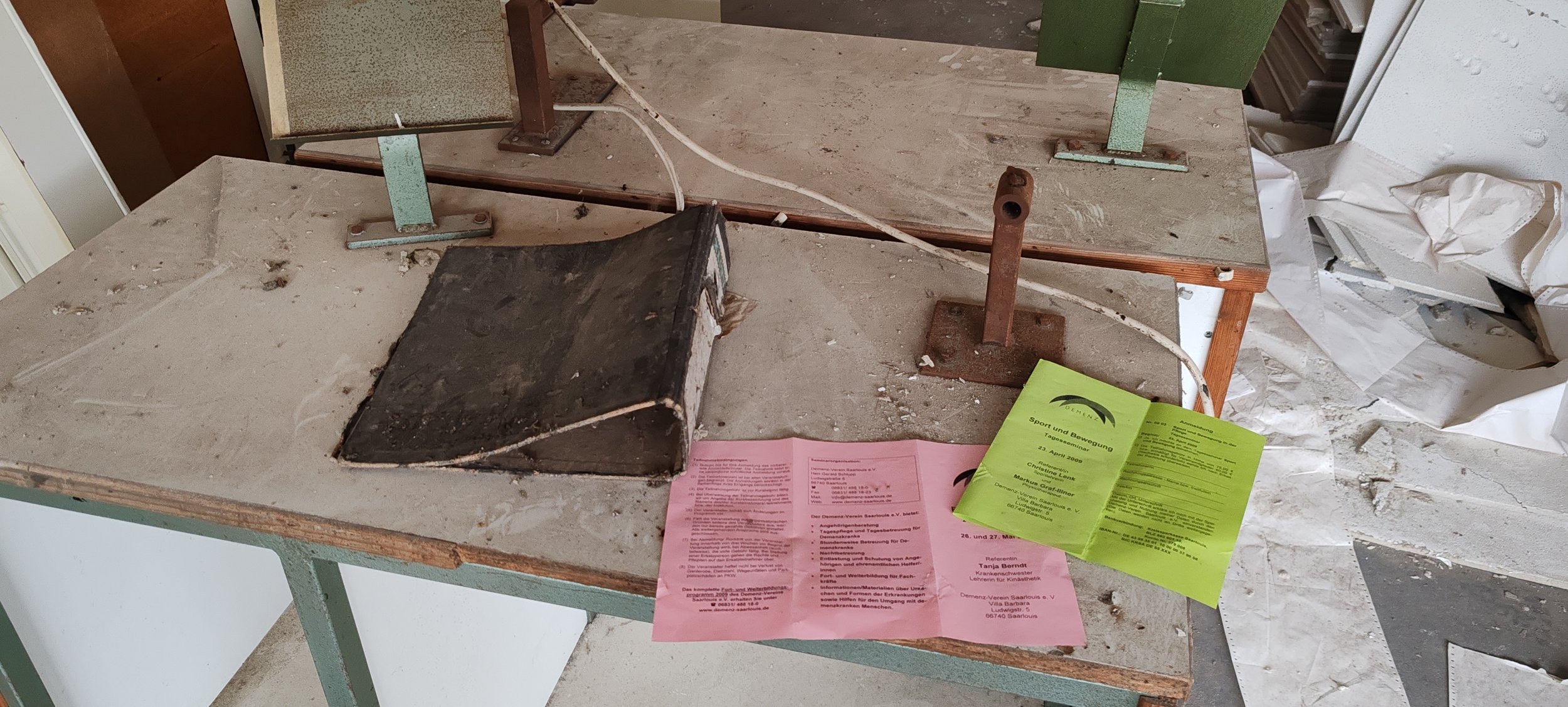 Ein alter, verstaubter Tisch mit Lego-Steinen und verschiedenen Papieren, darunter eine grüne und eine rosa Broschüre, in einem chaotischen Raum.