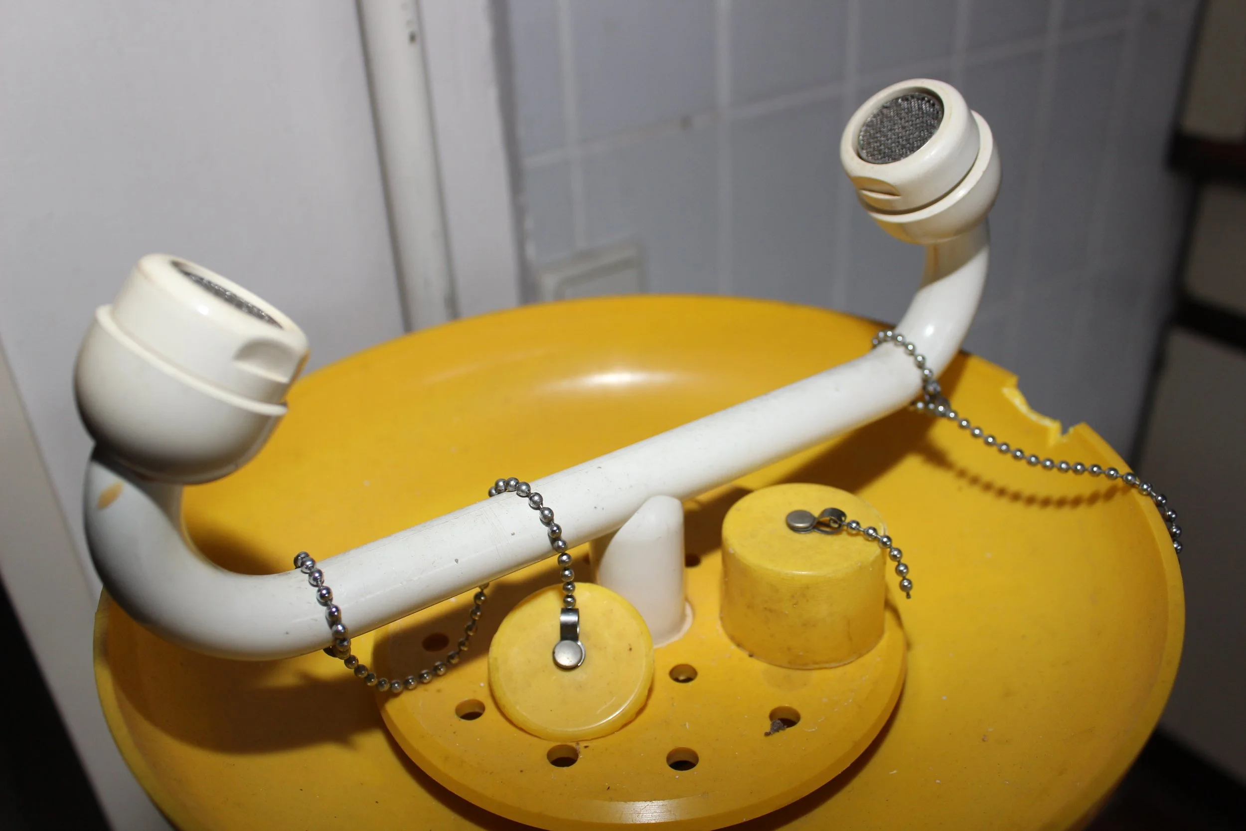 Ein altes, zerbrochenes gelbes Notruftelefon mit einem weißen Hörer und einer Kette, die den Hörer verbindet, liegt auf einer gelben Oberfläche.