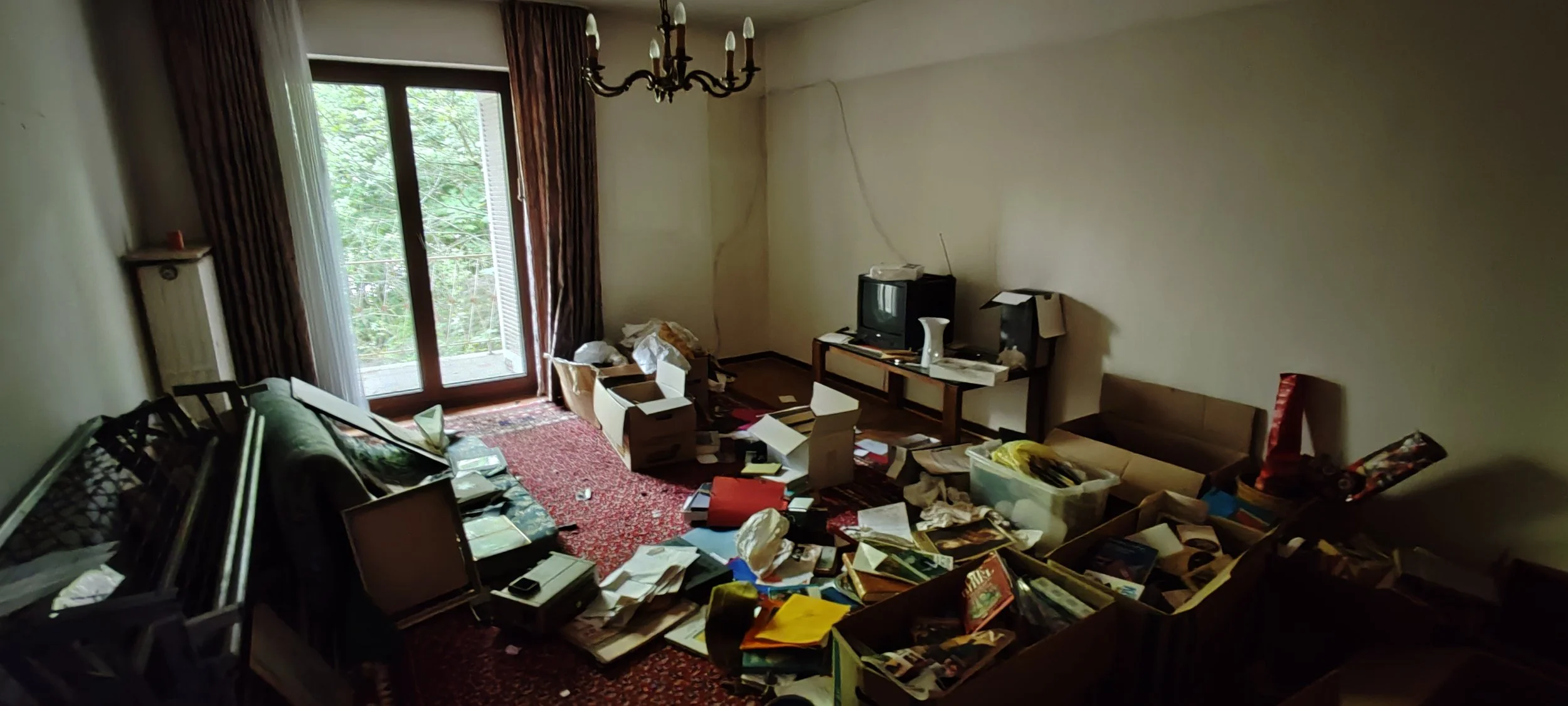 Un unordentliches Wohnzimmer mit Büchern, Papieren, Kartons und Möbeln, Fernseher, Balkon mit Gartenblick, und antiker Kronleuchter.
