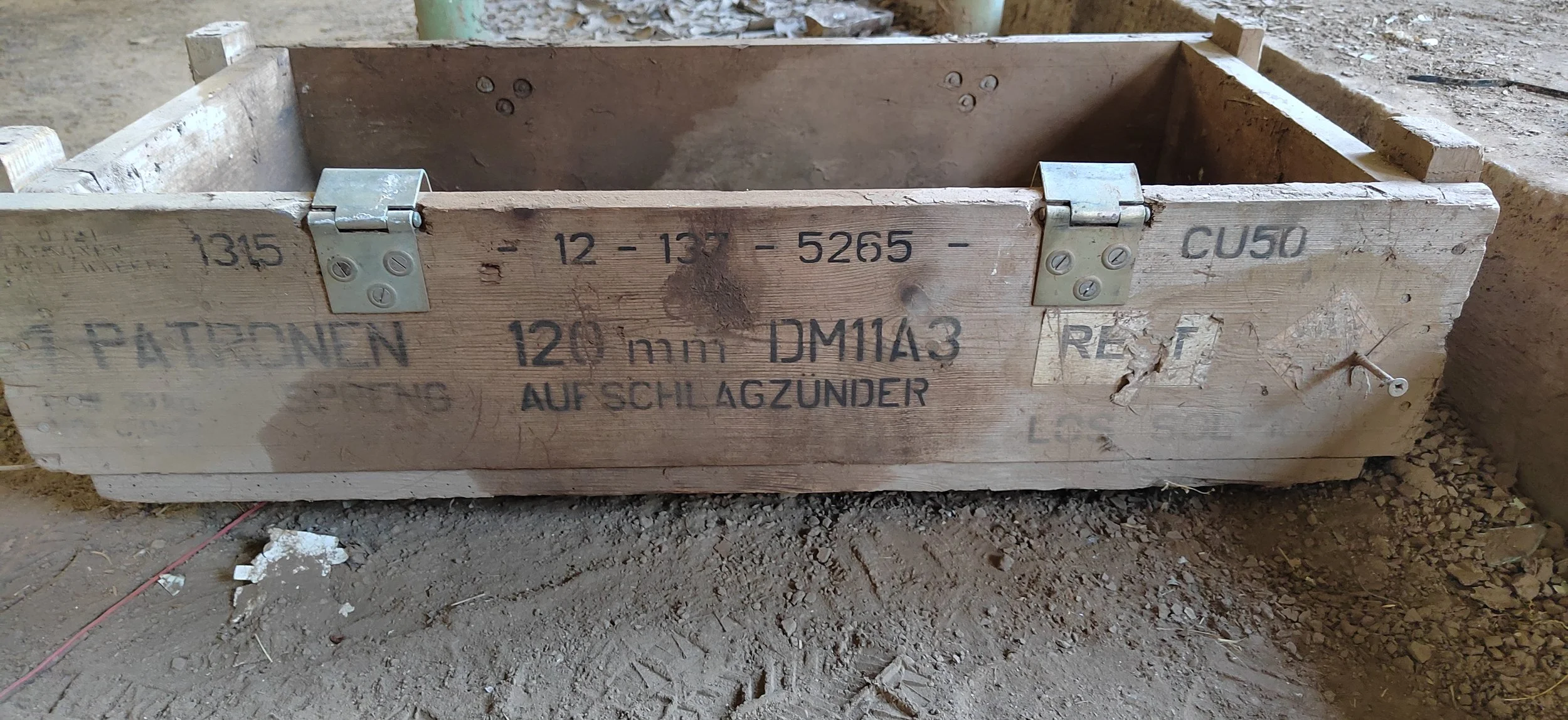 Holzkiste mit Metallbeschlägen, die auf einem Boden steht. Die Kiste hat deutsche Beschriftungen und einige Schrauben, sowie sichtbare Gebrauchsspuren.