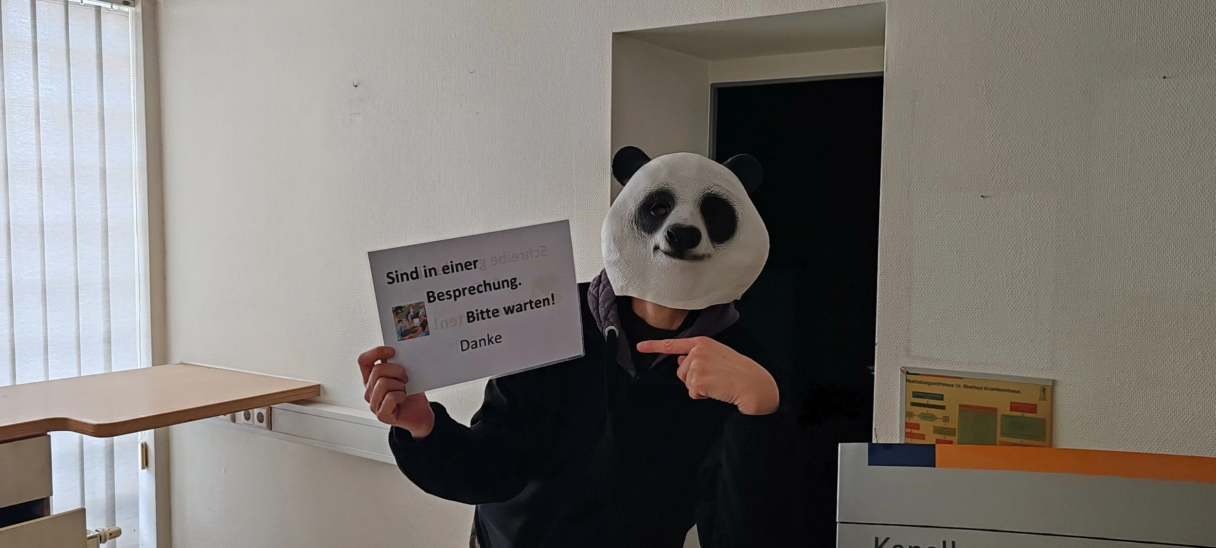 Person trägt eine Pandamaske und hält ein Schild mit der Aufschrift 'Sind in einer Besprechung. Bitte warten! Danke'.
