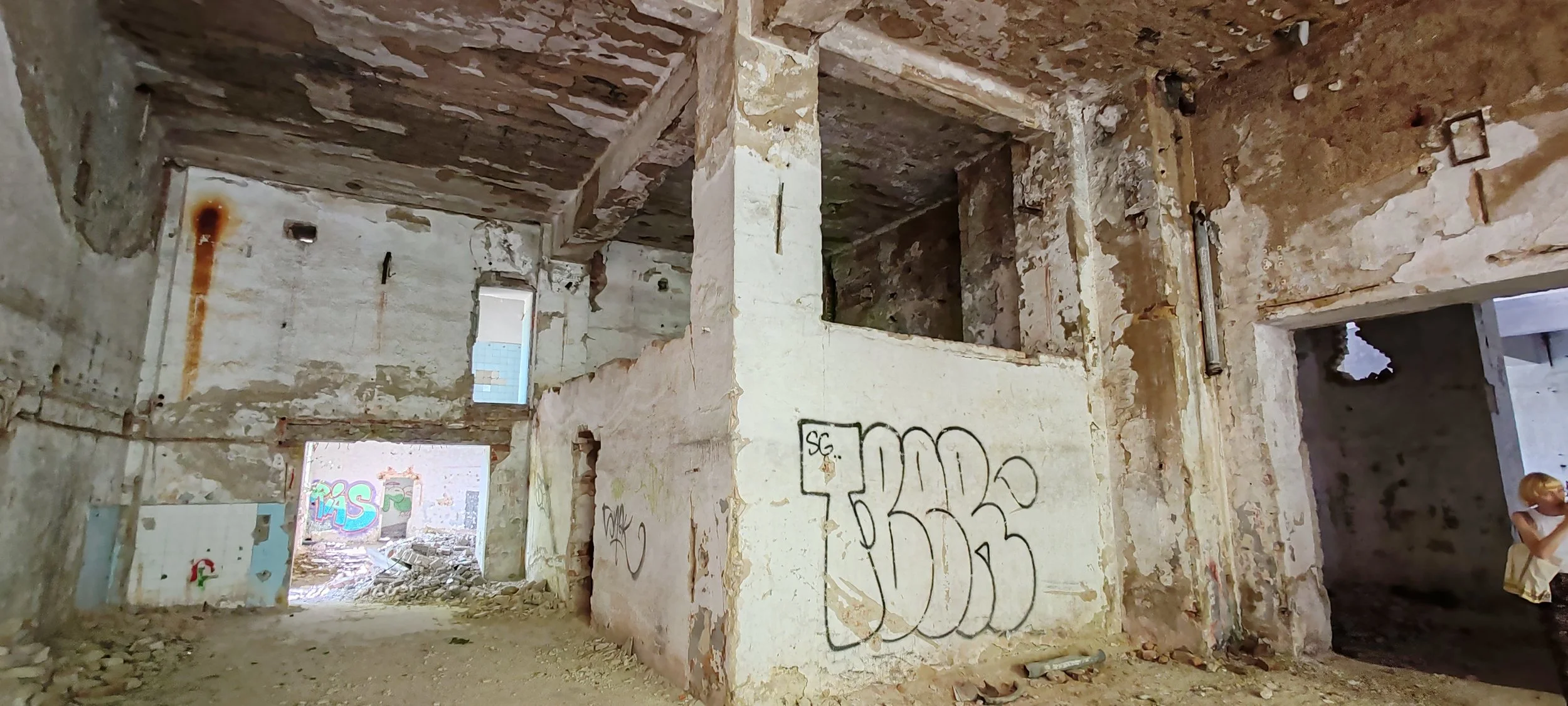 Verlassene, verfallene Indoor-Struktur mit graffiti-beschmückten Wänden, Bruchstellen und zerbrochenen Fenstern, darunter eine Person mit Sonnenbrille und goldener Perücke, die eine Wasserflasche hält.