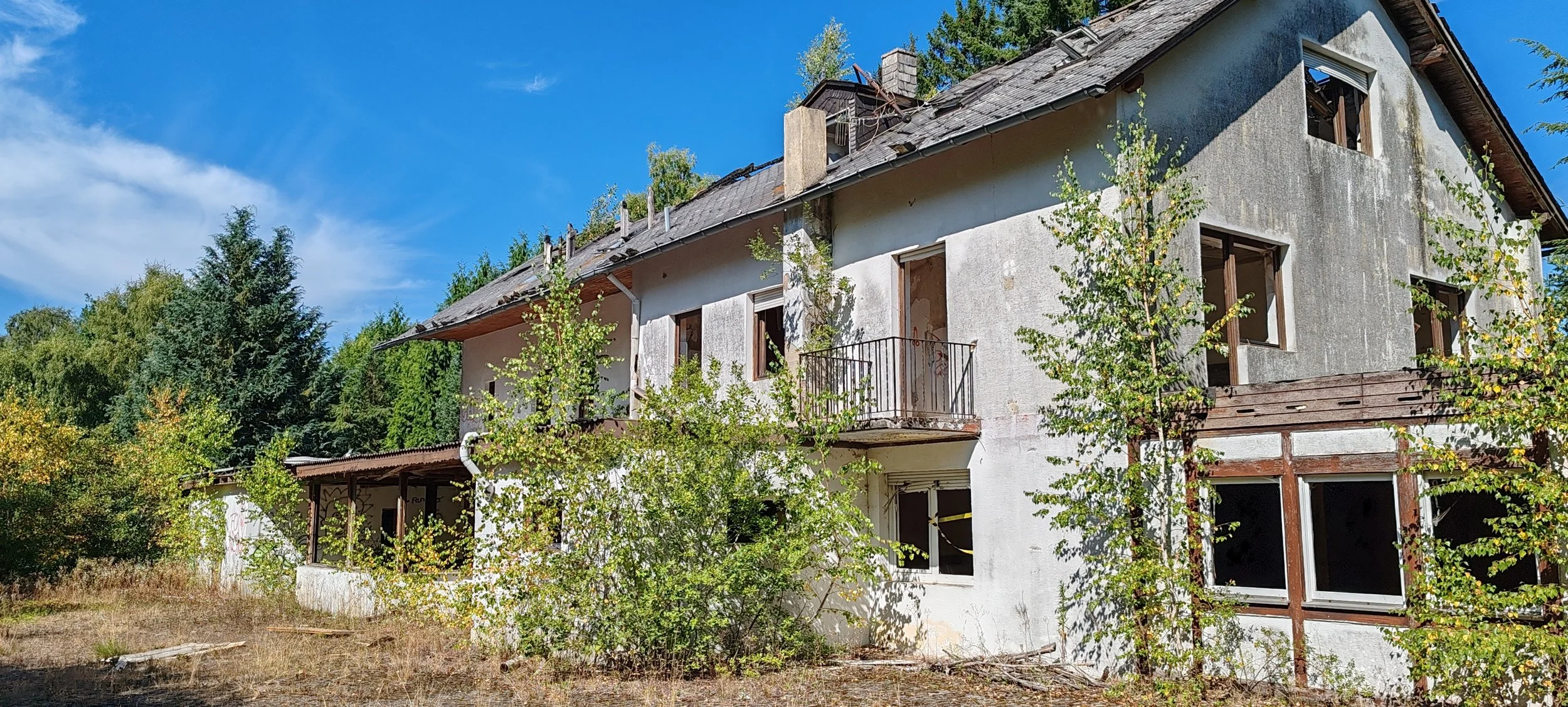 Verfallenes, zweistöckiges Haus mit beschädigten Fenstern und überwucherten Bäumen im Vordergrund, umgeben von Bäumen unter einem blauen Himmel.