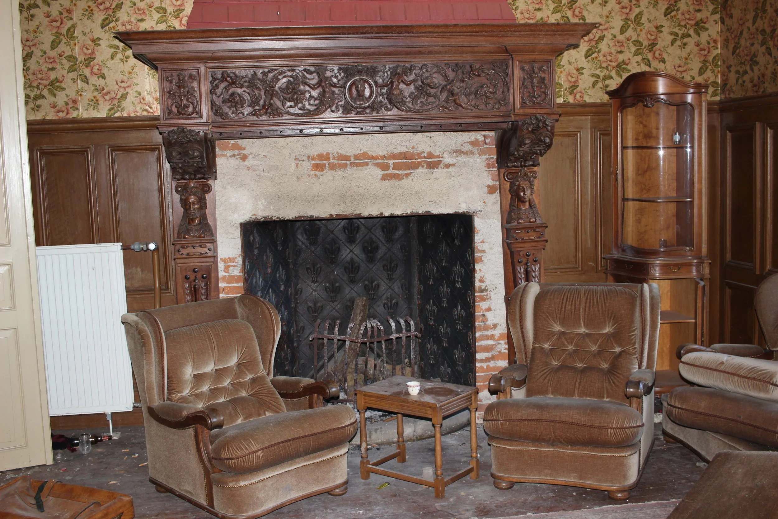 Alte Wohnzimmer mit antikem Kamin und braunen Sessel, Holzwandverkleidung und floral bestickter Tapete.
