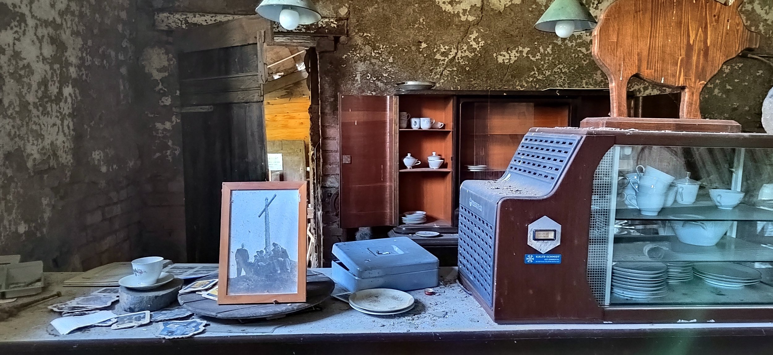 Veralteter, unordentlicher Raum mit zerbrochenen Geschirr, Staub und alten Gegenständen, darunter eine Glasscheibe, ein Mikrowellengerät, eine offene Holzschrank, eine Glaskühlschrank mit Porzellan.