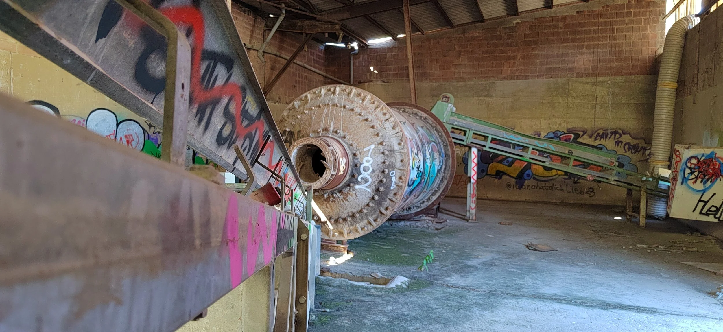 Ein verlassener und mit Graffiti beschmierter Kellerraum mit einem alten, rostigen mechanischen Teil, Wasserpfützen auf dem Boden und einer auffangenden Rampe oder Förderband.