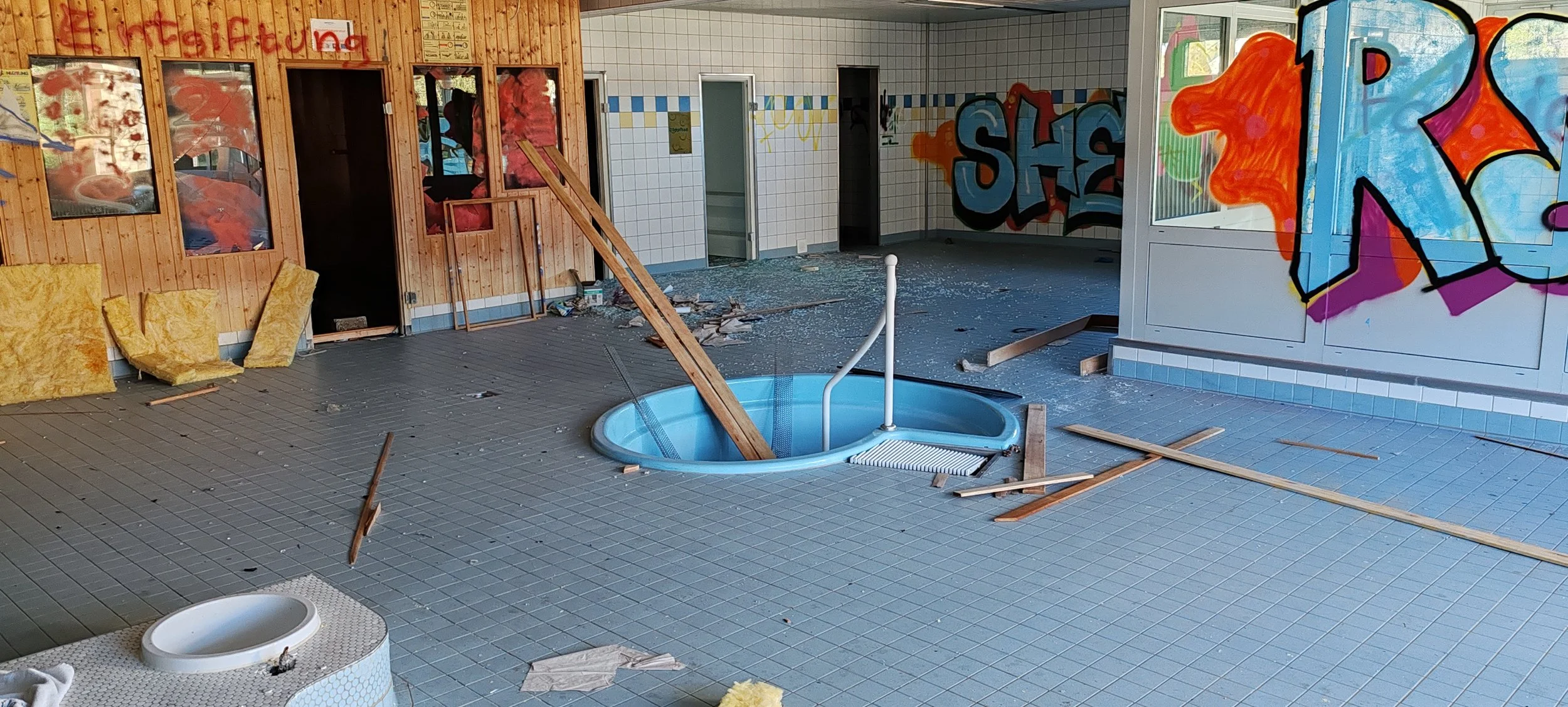 Innenraum eines Schwimmbads unter Renovierung mit Baugerüsten, Baumaterialien, zerbrochenem Glas und Graffiti an den Wänden.