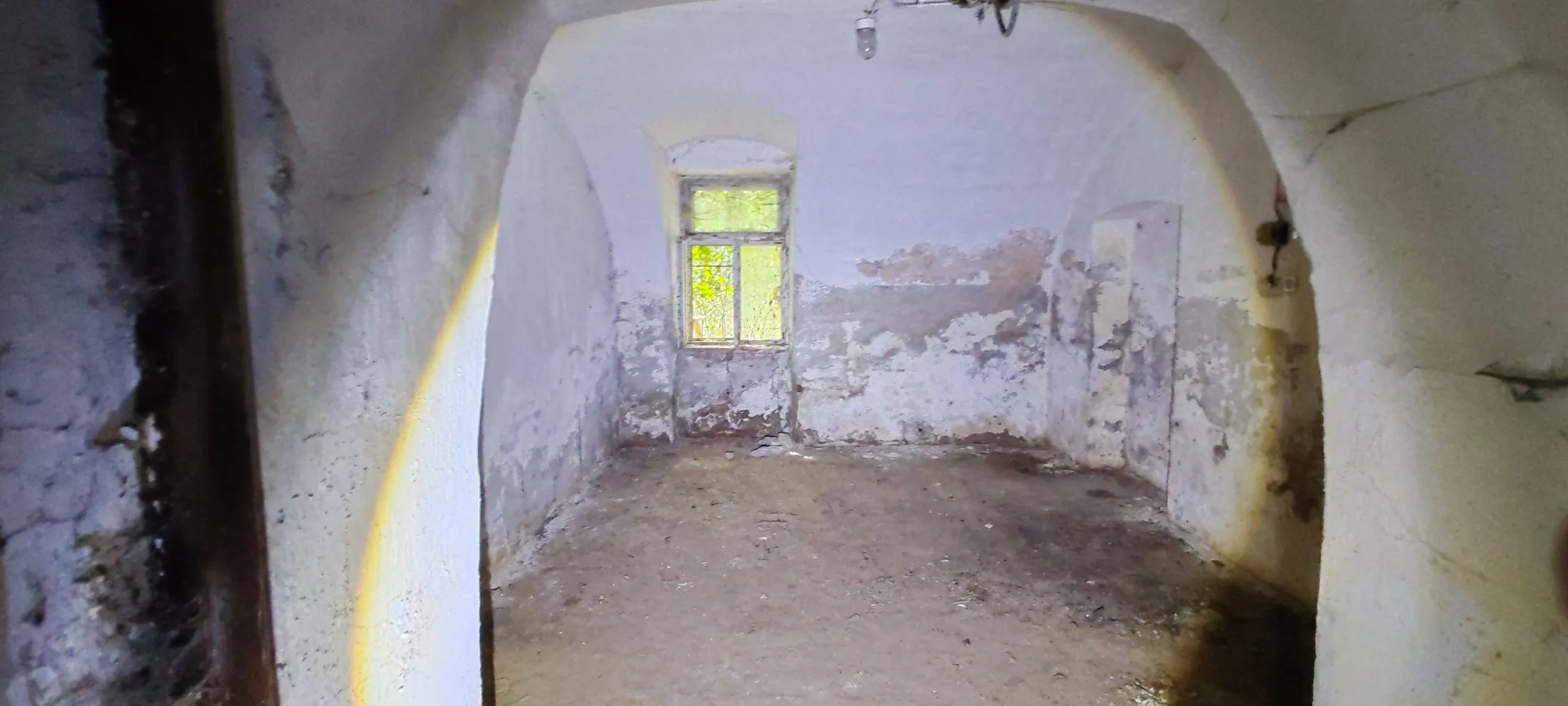 Ein verlassener, alter Raum mit verputzten, beschädigten Wänden, einer kleinen Fensteröffnung, Staub und Schmutz auf dem Boden.