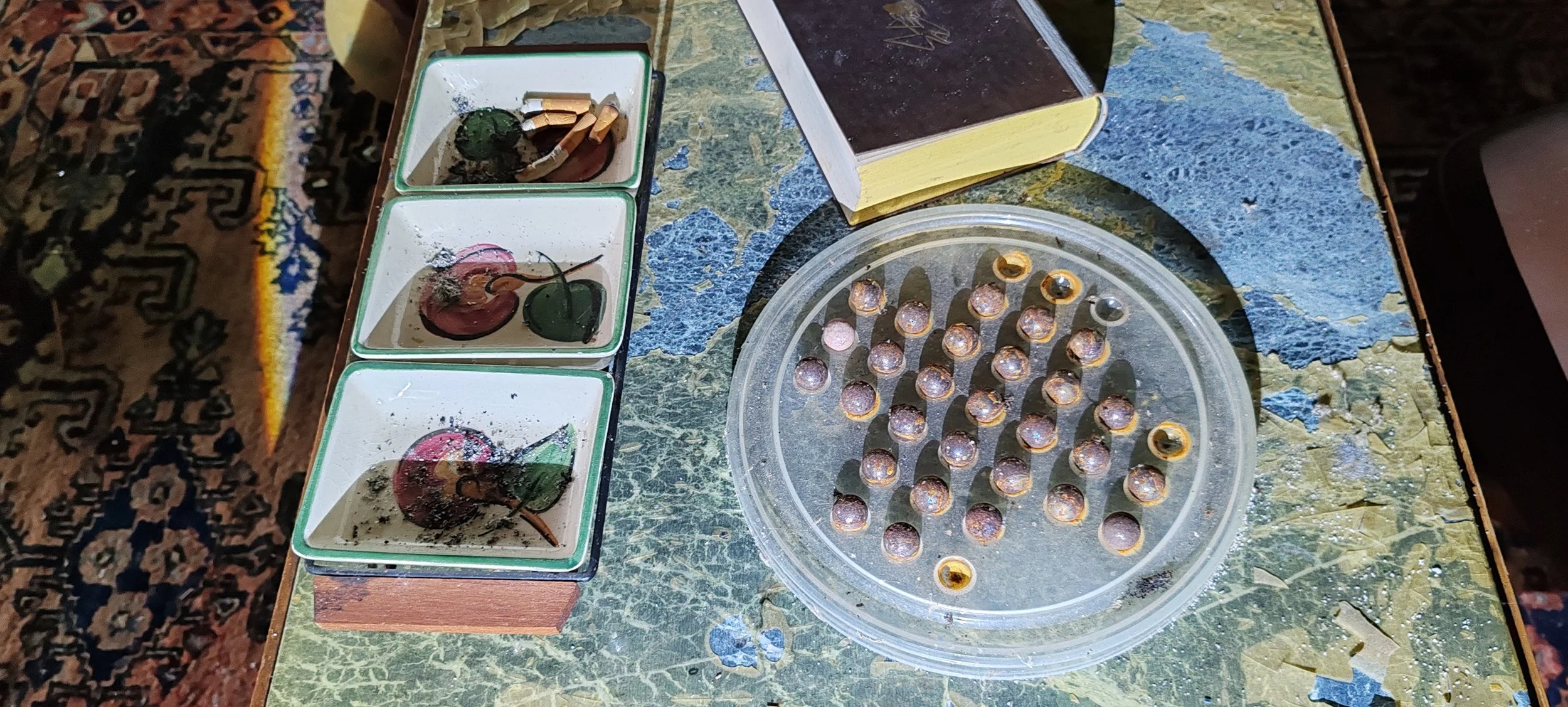 Dreie Unterteller mit krümeligen, verbrannten Keksen, einer runden durchsichtigen Vorratsdose mit zahlreichen unregelmäßig geformten, glänzenden, metallisch schimmernden Kugeln, ein offenes Buch und eine bunte, gemusterte Tischdecke.