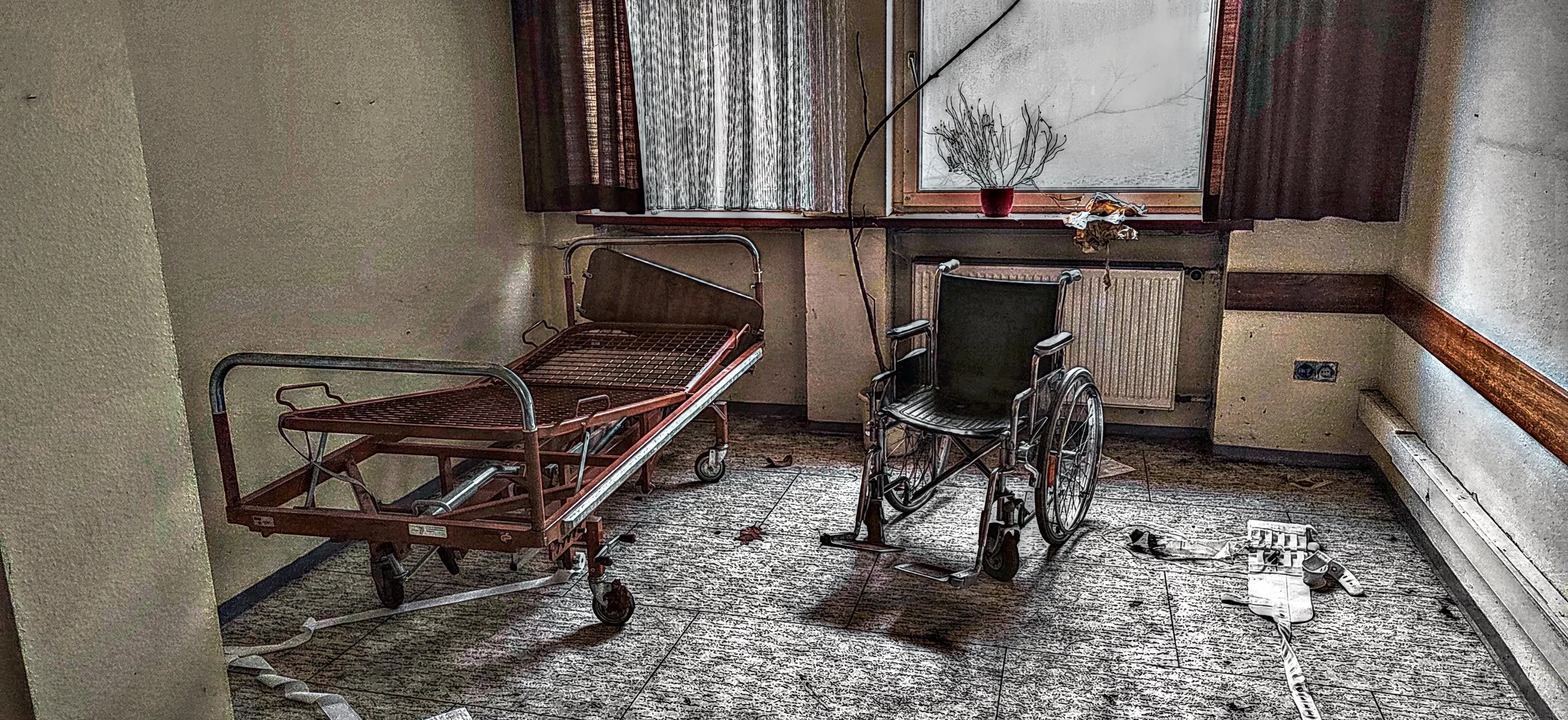 Ein verlassener, verstaubter Raum mit einem Krankenhausbett und einem Rollstuhl, Staub und Schmutz auf dem Boden, zerbrochene Gegenstände, verstaubte Fenster mit alten Vorhängen.