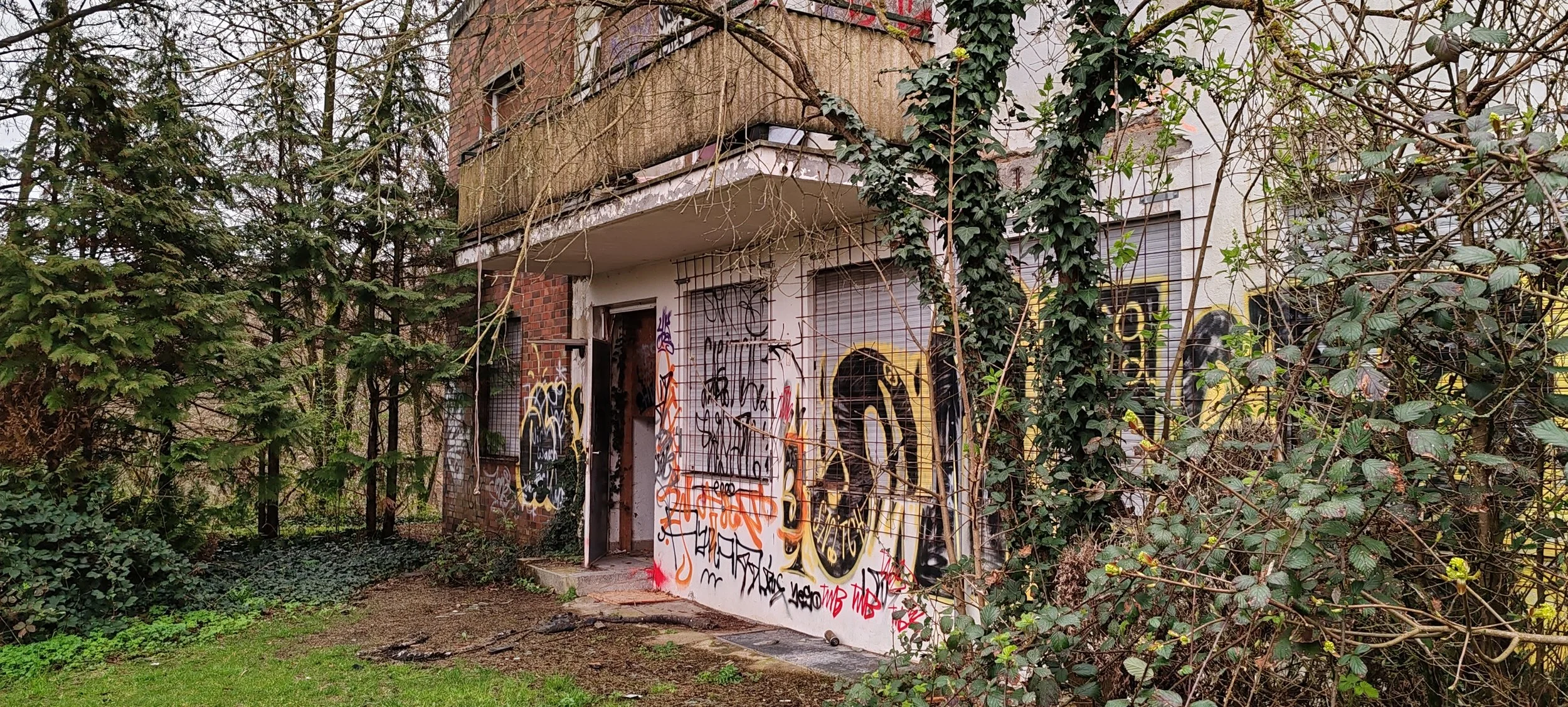 Verfallene Gebäude mit Graffiti an den Wänden, umgeben von Bewuchs und Bäumen.