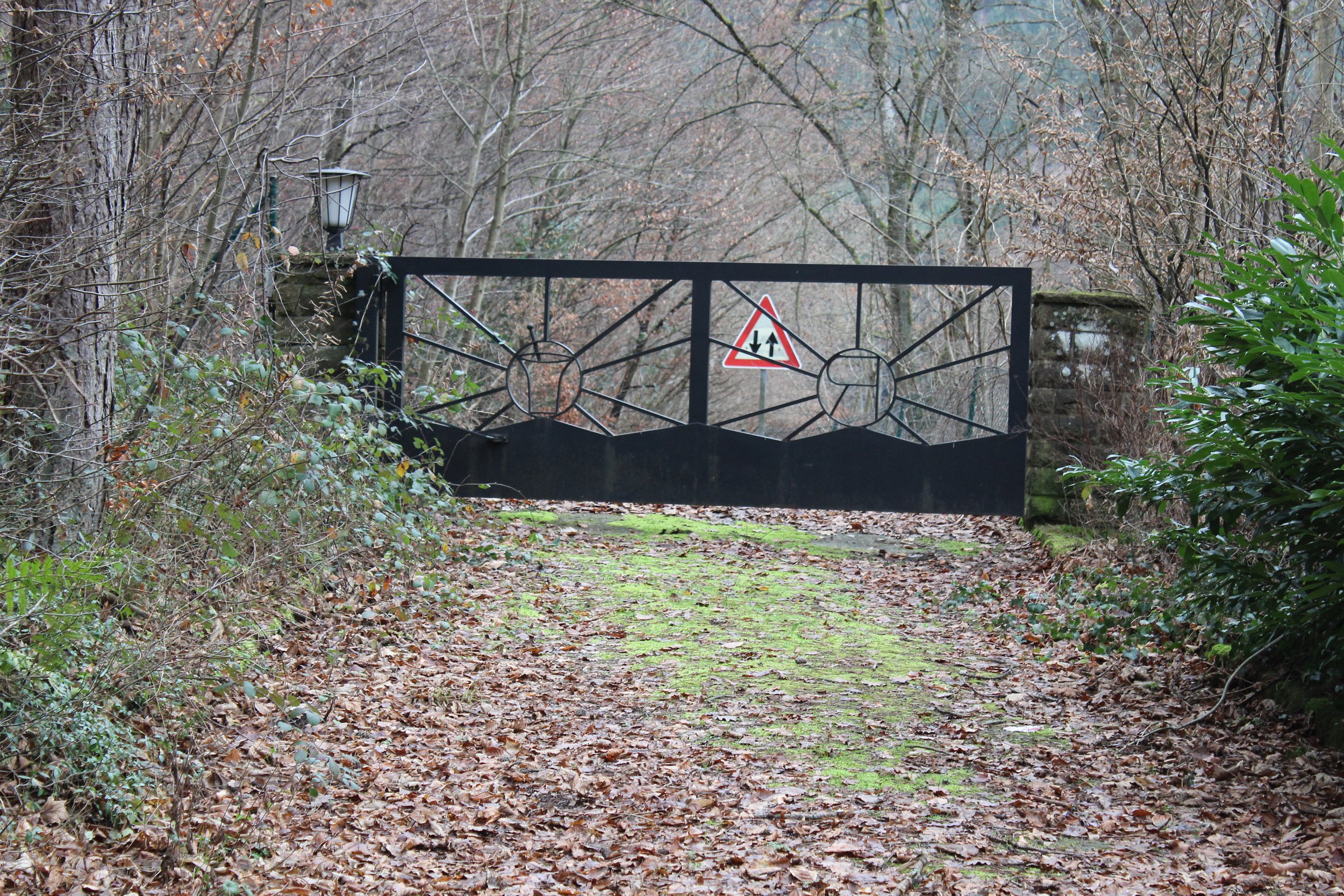 Ein verschlossener Gitterzaun mit einem Warnhinweis, umgeben von Bäumen und Büschen, auf einem abgedeckten Weg im Wald.