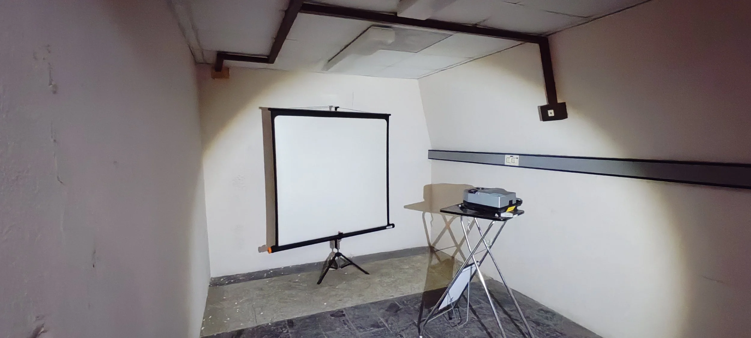 Ein leeres, kleines Zimmer mit weißen Wänden und dunklem Boden. Im Raum befinden sich eine Projektorleinwand auf einem Ständer und ein Tisch mit einem Beamer. Die Decke hat eine einfache Lichtquelle.