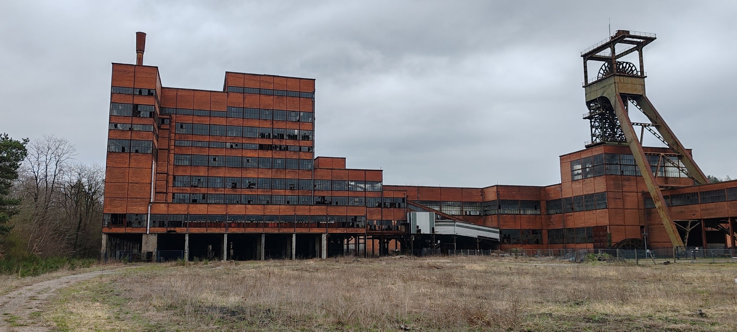 Verlassene Industrieanlage mit großen roten Backsteingebäuden und einem Förderturm, umgeben von einem unbebauten, verfallenen Grundstück, bei wolkigem Himmel.
