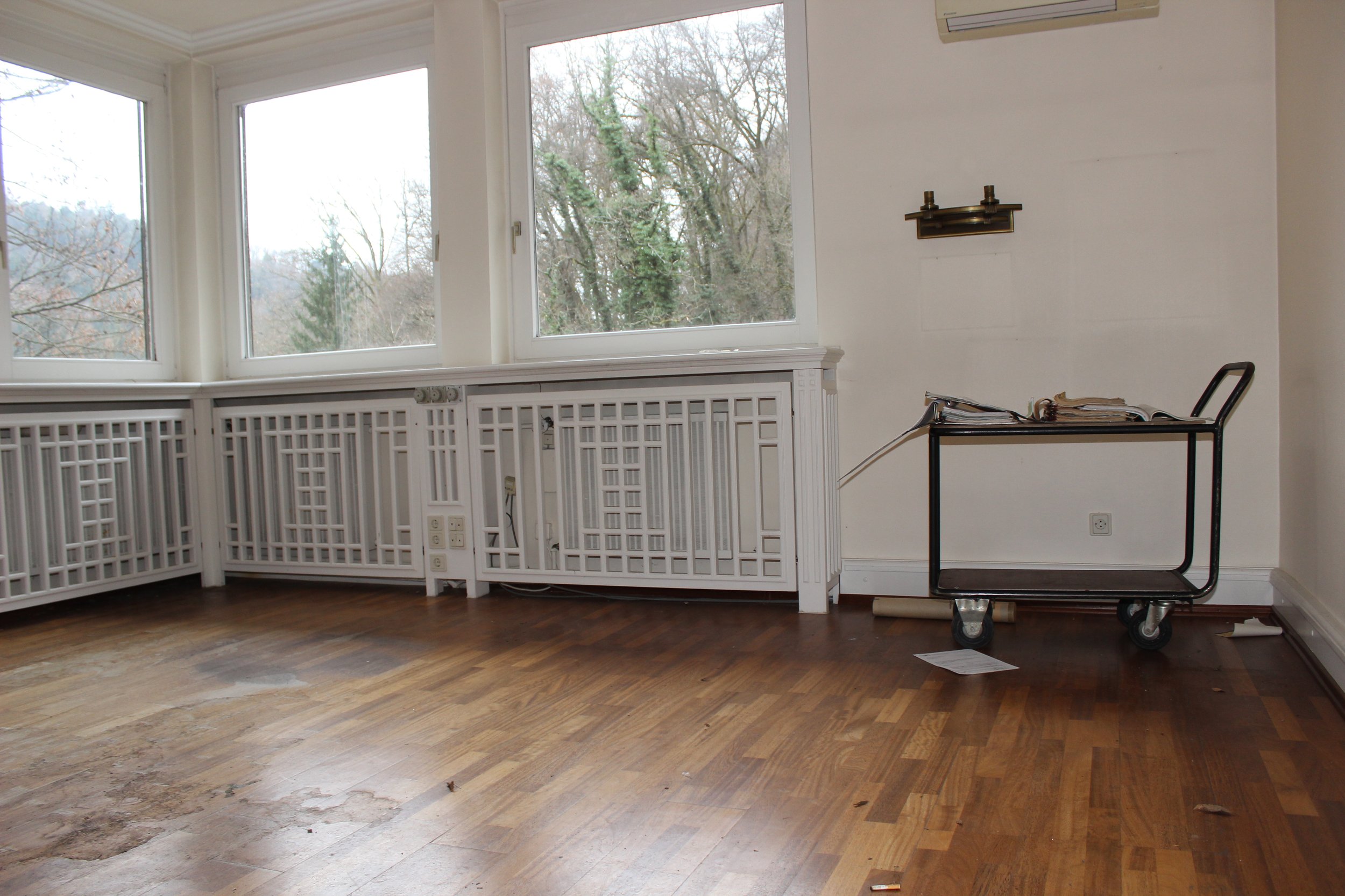 Ein unaufgeräumtes, hell erleuchtetes Wohnzimmer mit großen Fenstern, Holzboden und einem serveiertisch auf Rollen, der mit Papieren und Büchern bedeckt ist.