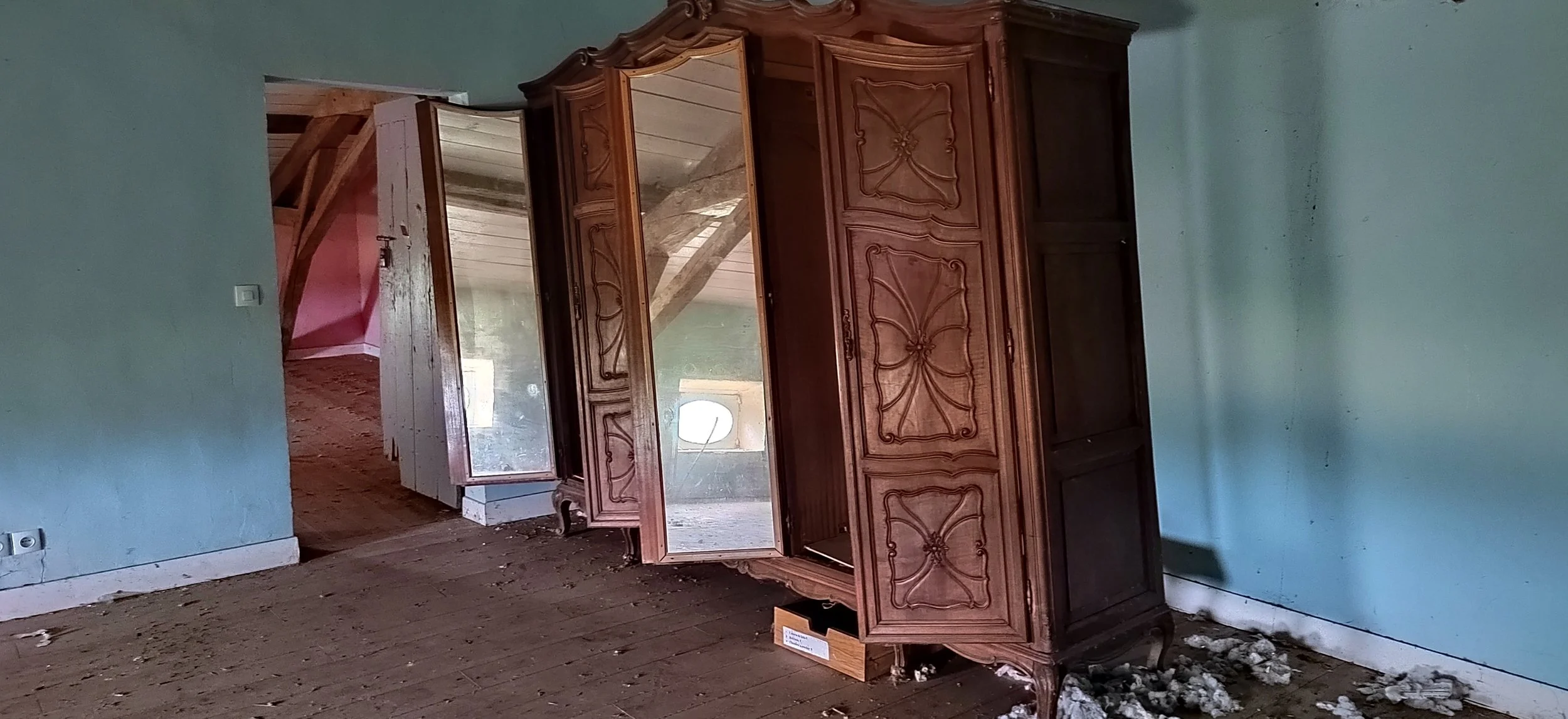 Ein antiker Holzschrank in einem verlassenen Raum mit blauen Wänden und Holzboden, durch den ein angrenzender Raum sichtbar ist.