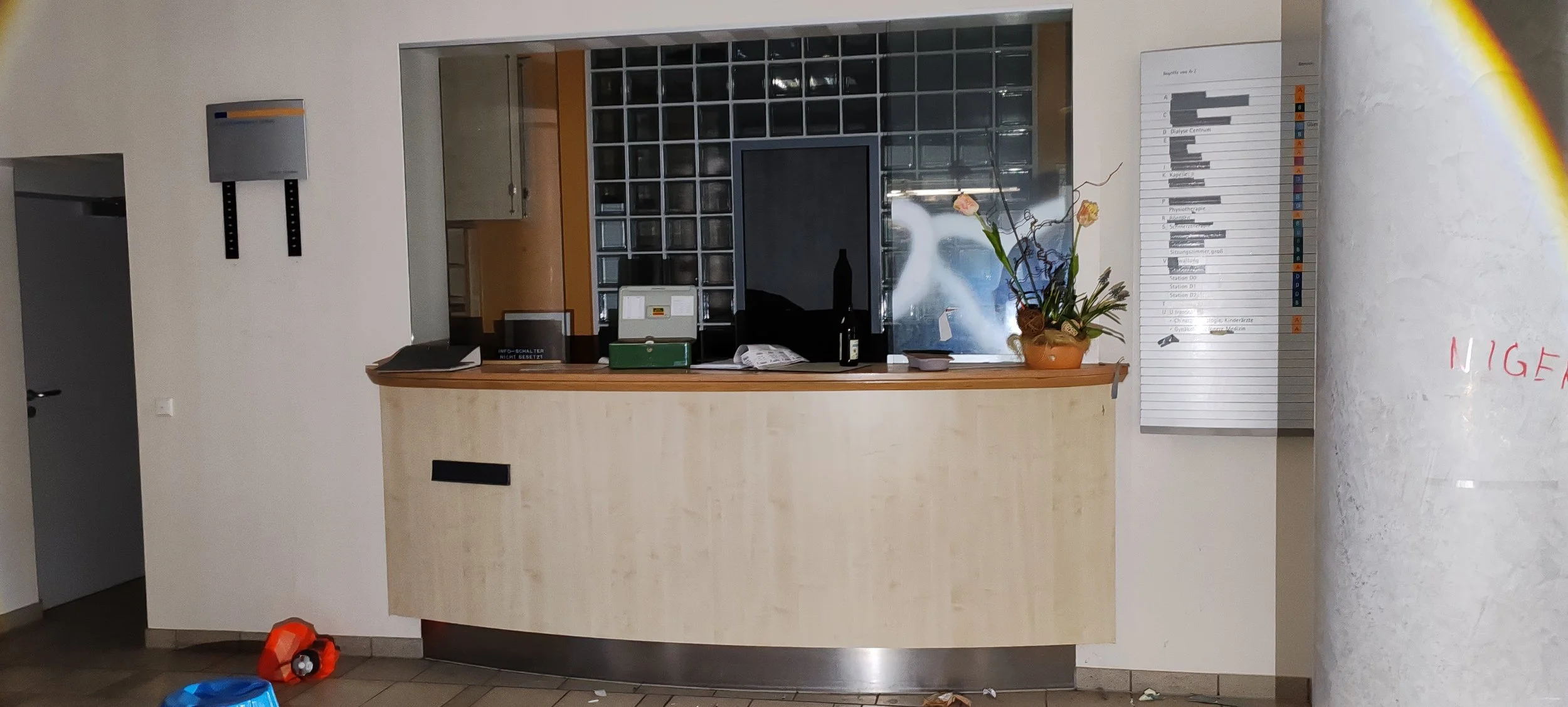Blick auf einen Empfangstresen in einem öffentlichen Bereich mit einem Blumenstrauß, einem schwarzen Fernseher, einem Getränke, und hinter einem Fenster im Hintergrund eine Küche. An der Wand rechts ein schwarzer Plan mit Stundenangaben, auch an der 