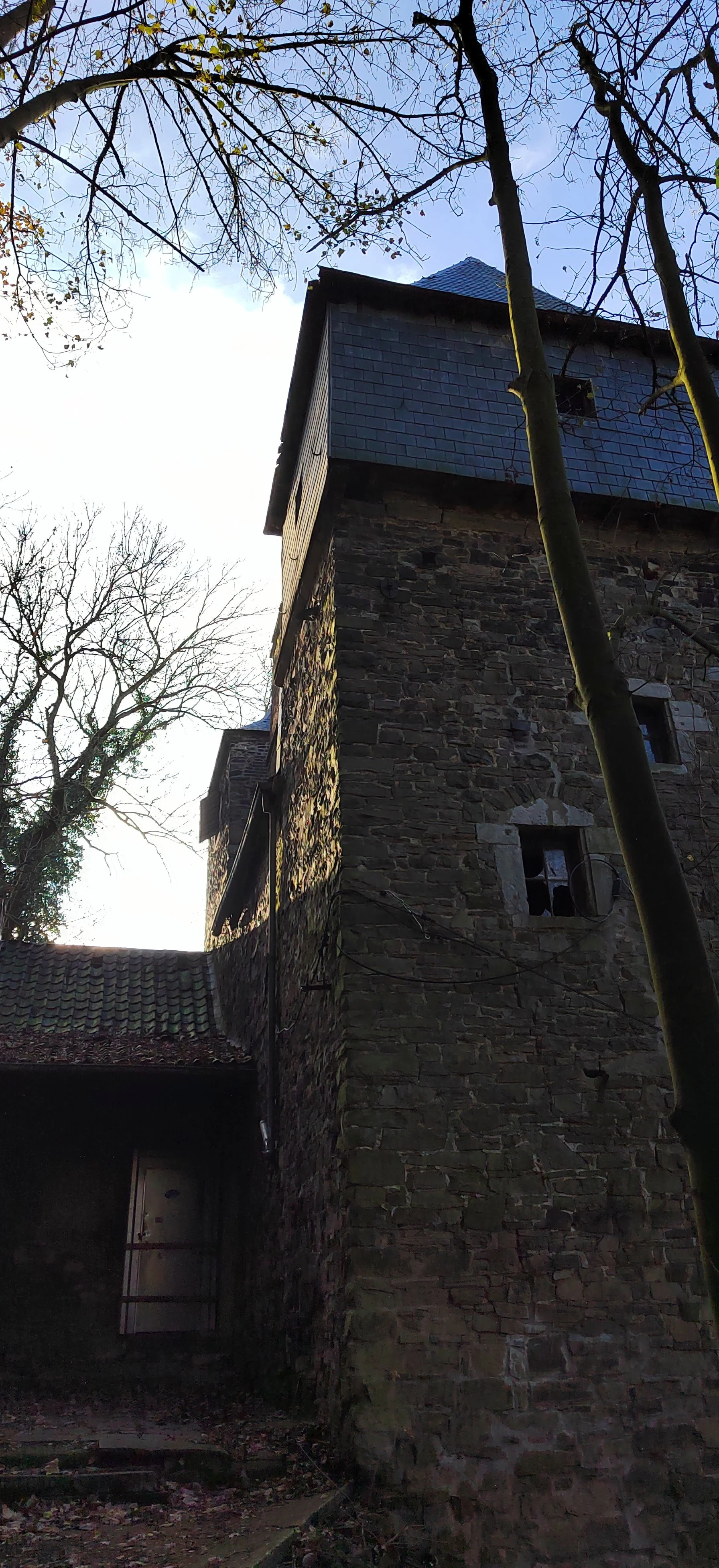 Ein altes Steingebäude mit kleinen Fensteröffnungen und einem steinernen Turm, umgeben von Bäumen mit kahl werdenden Ästen, bei Sonnenuntergang oder -aufgang.