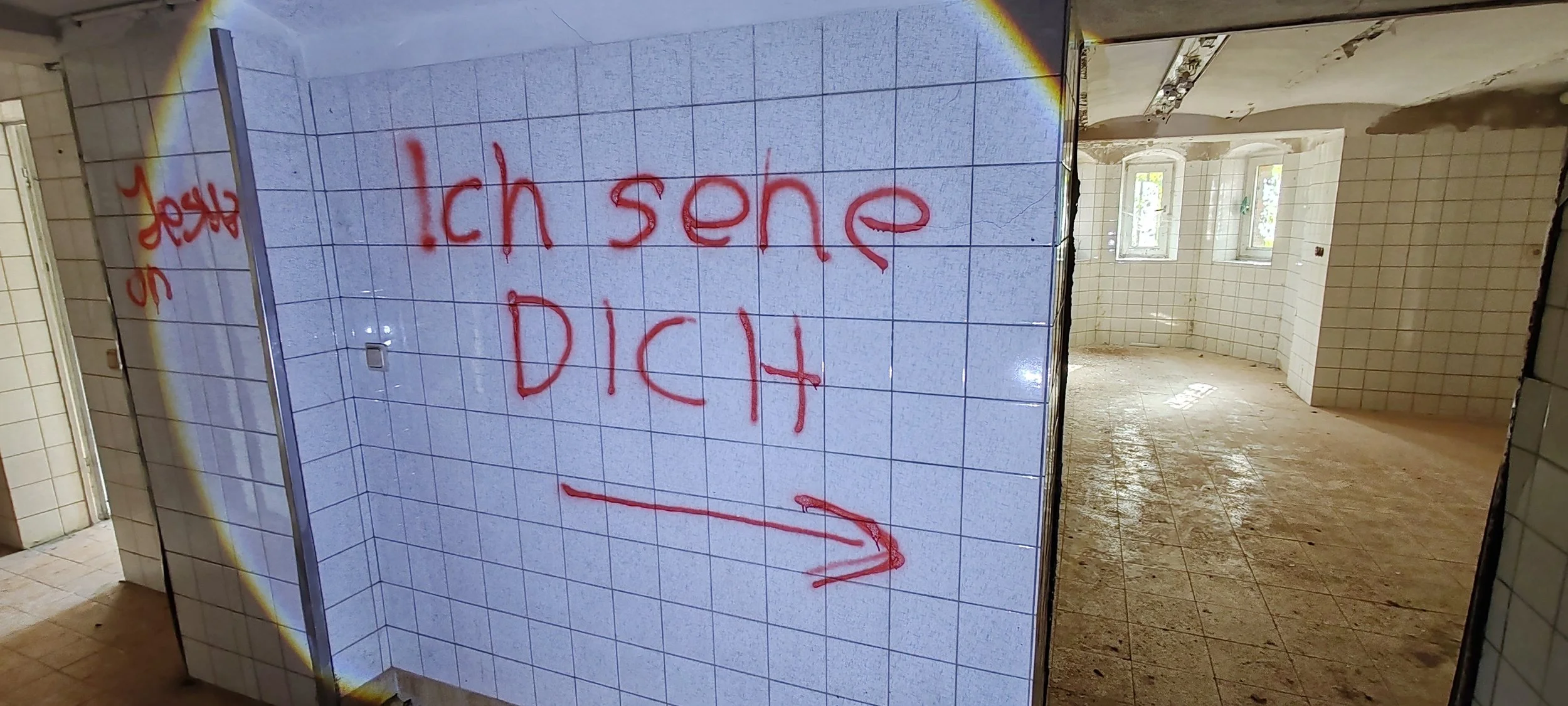 Verlassener Raum mit weißen Fliesen, Auswahl an Fenstern und rot geschriebenem Graffiti auf einer Wand, das sagt: 'Ich sende DICH' mit einem Pfeil nach rechts.