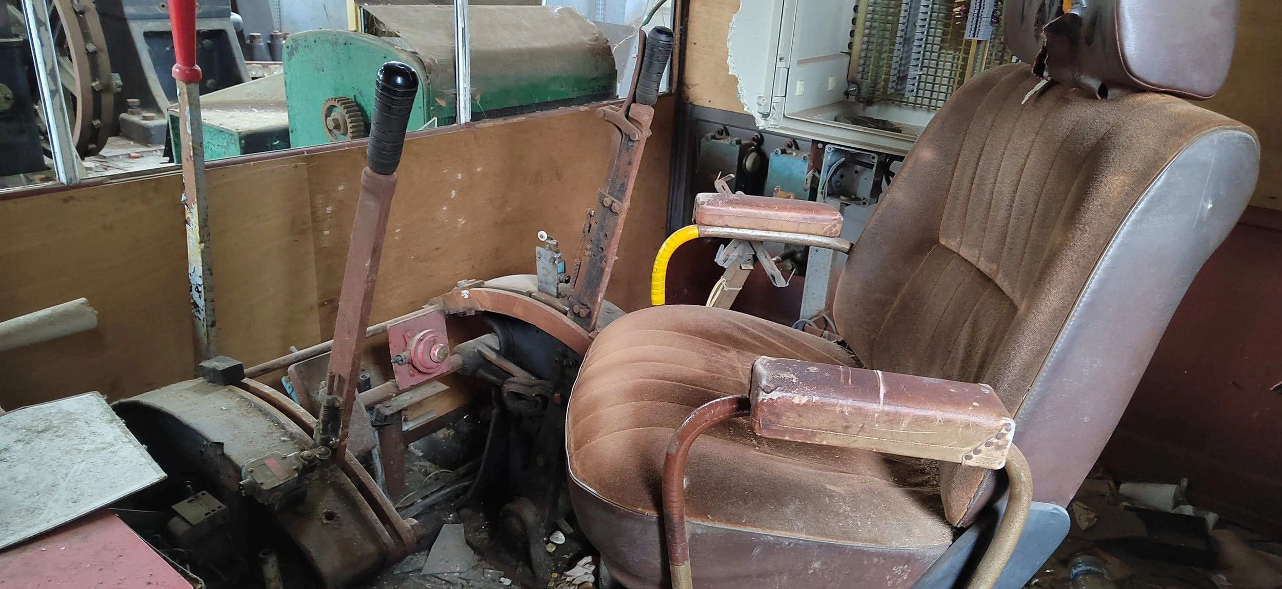 Ein alter, verstaubter Sitz mit Armlehnen in einem Werkstatt- oder Schulungsraum, umgeben von Werkzeugen und Maschinen.