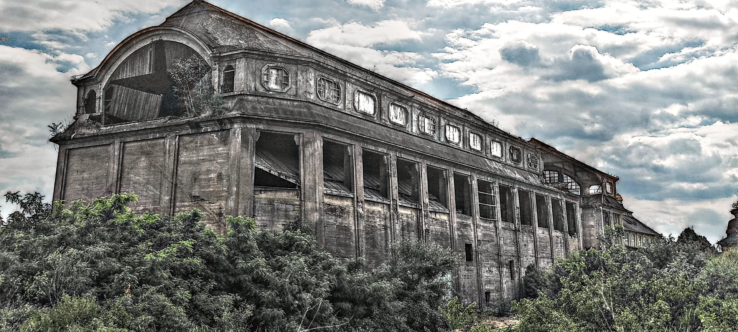 Verfallenes mehrstöckiges altes Gebäude mit zerbrochenen Fenstern, umgeben von Büschen und Bäumen, unter bewölktem Himmel.