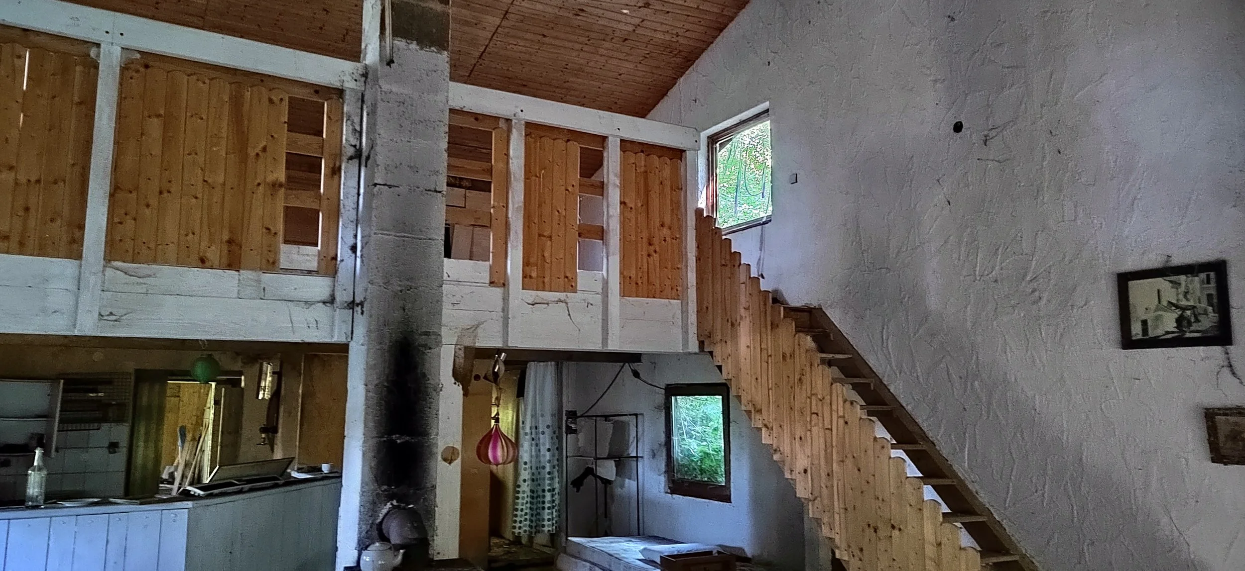 Innenansicht eines Hauses mit einer Holztreppe, weißen Wänden mit Strukturoberfläche, Fenstern, und einer kleinen Küche im unteren Bereich, dekoriert mit Lampen und Bildern.
