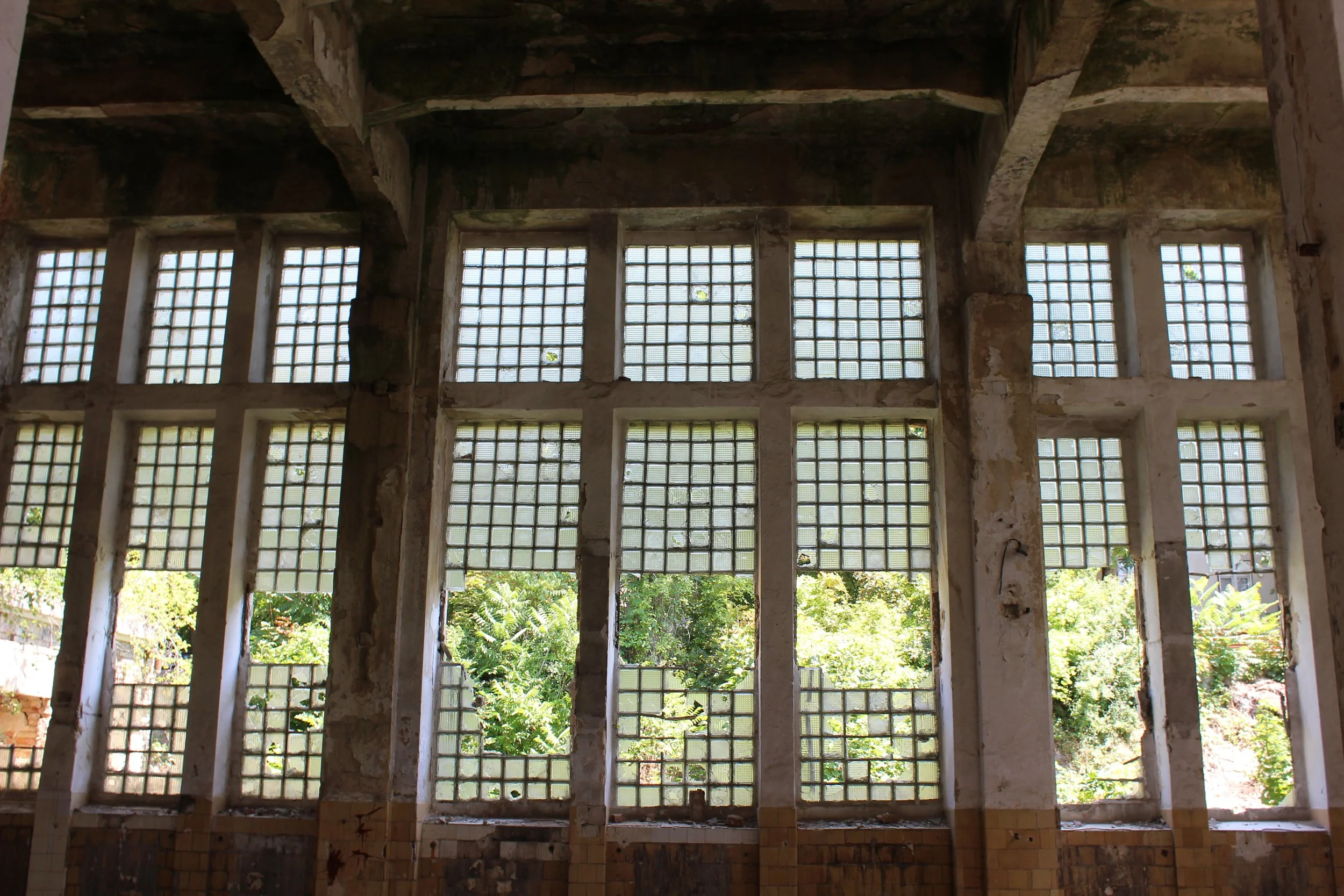 Bunte Fenster mit Gitter in einem alten Gebäude mit Blick ins Freie, umgeben von grünen Bäumen.